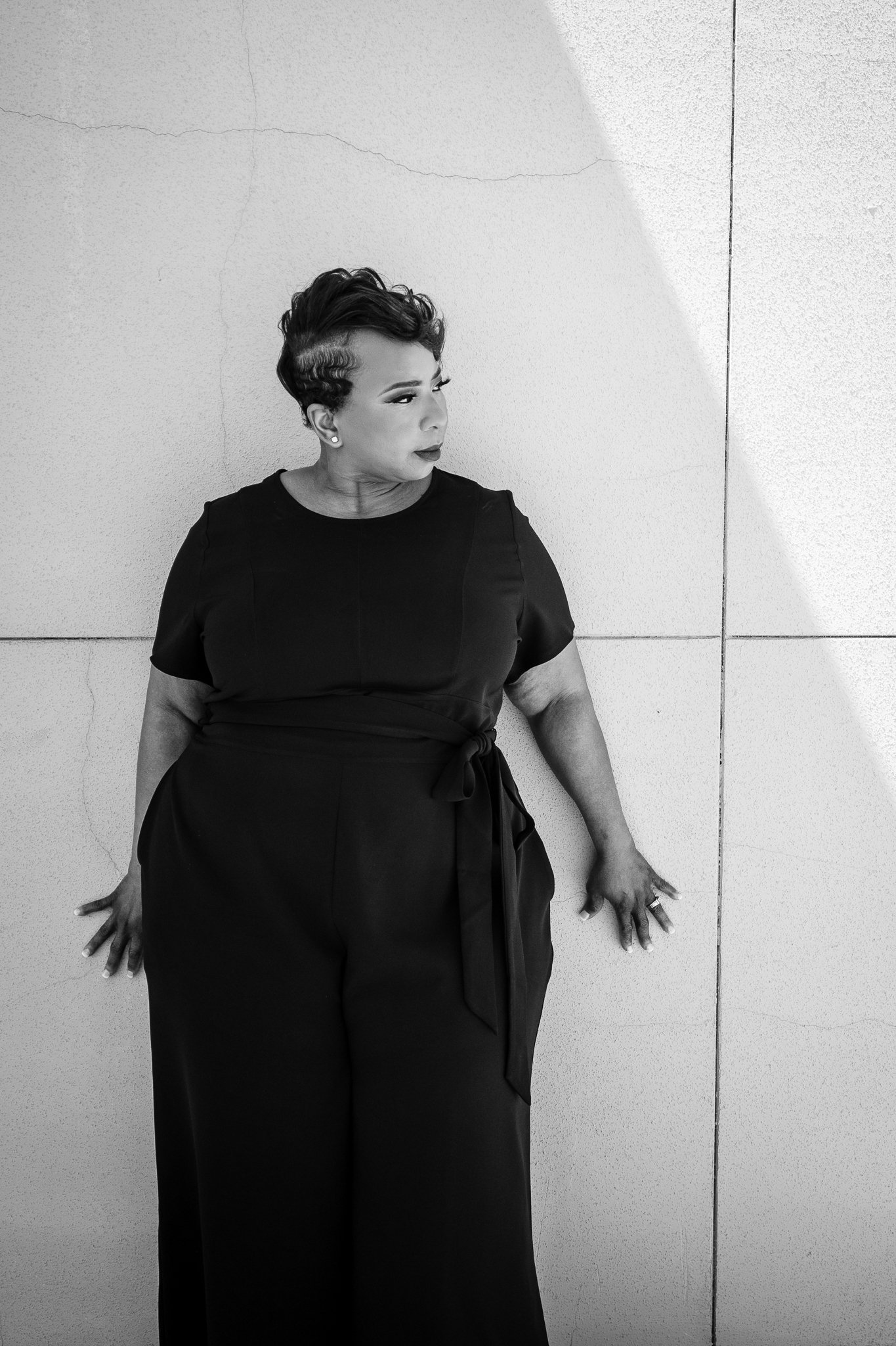 Black and white photograph of a woman leaning against a cracked wall, looking to her left with her hair styled in vintage waves, wearing a black dress with a bow on the side.