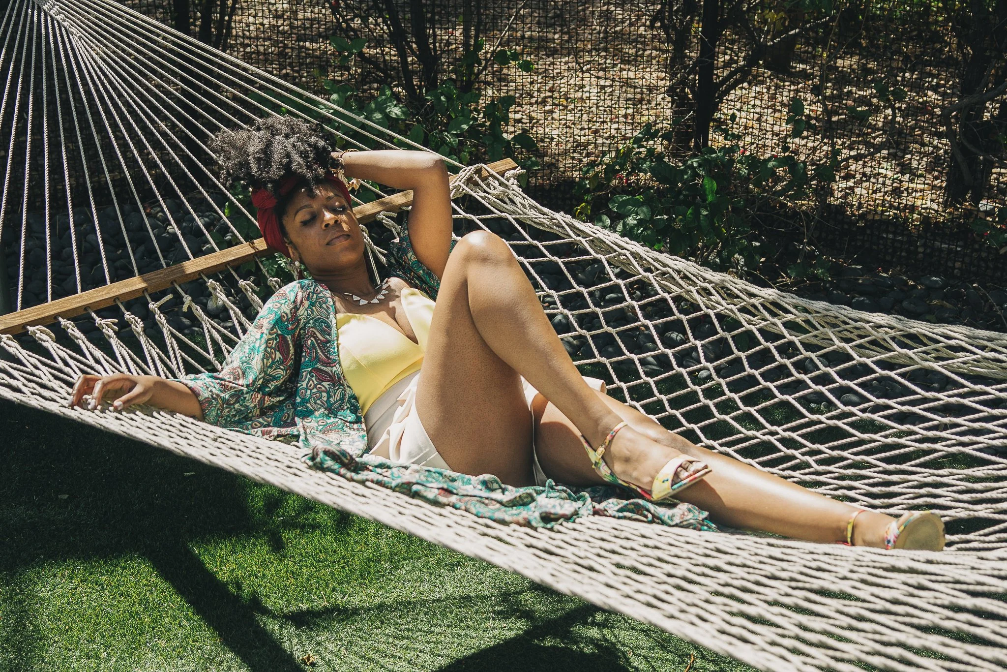 Woman lying on a hammock outdoors, relaxing with her eyes closed, wearing a yellow top, colorful patterned jacket, white shorts, and floral high-heeled sandals, surrounded by greenery and a wire fence.