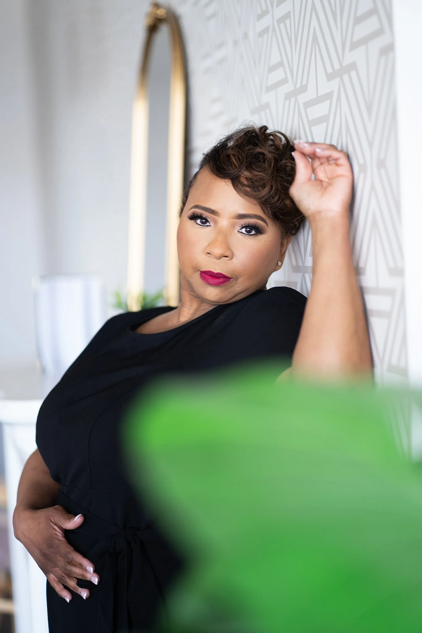 A woman with short curly hair, wearing a black top, posing indoors near a patterned wall and mirror, with a blurred green plant in the foreground.
