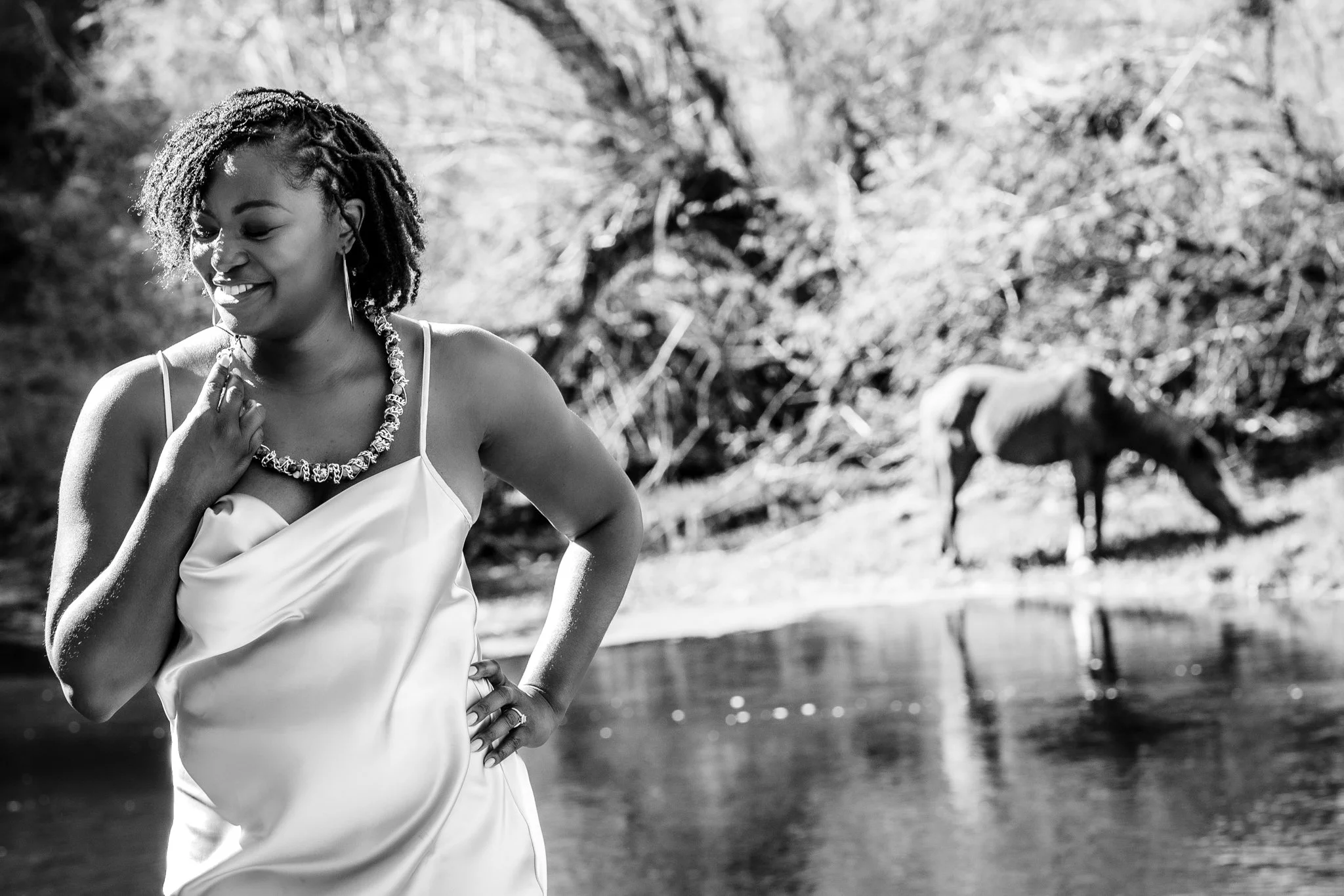 A woman with short curly hair wearing a satin dress and necklace, smiling by a river with a horse in the background.