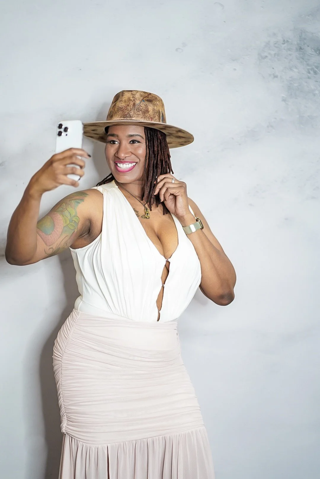 A woman taking a selfie, wearing a wide-brimmed hat, white top, and a light-colored skirt, standing against a plain wall.