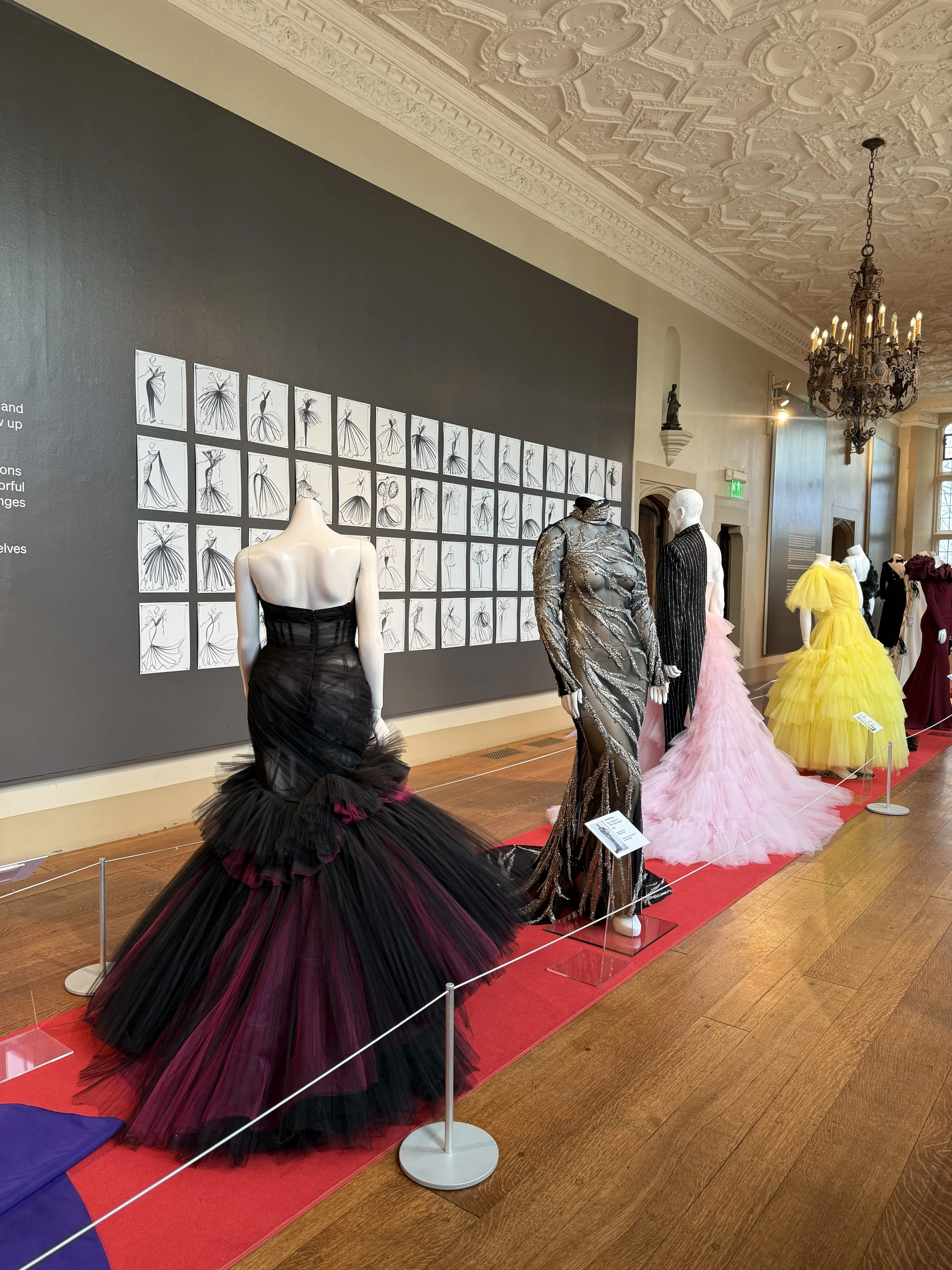 a collection of red carpet gowns designed by Christian Siriano on display at Branch Museum of Design, a gallery wall of drawn dress sketches in the background