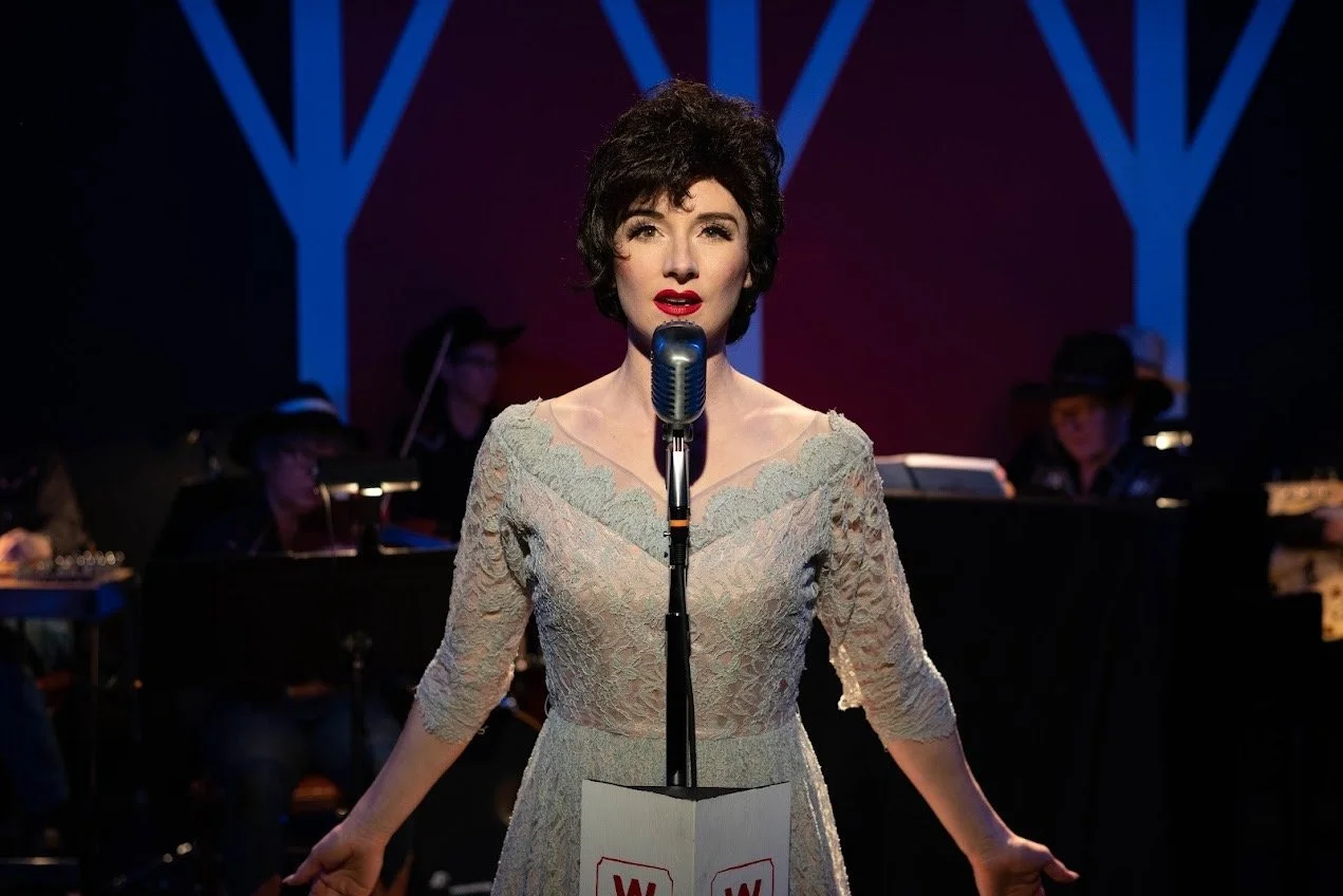  Abakis as Patsy Cline standing on stage in front of a band wearing a vintage blue dress with her hands down by her side. Image by Mel Carter. 