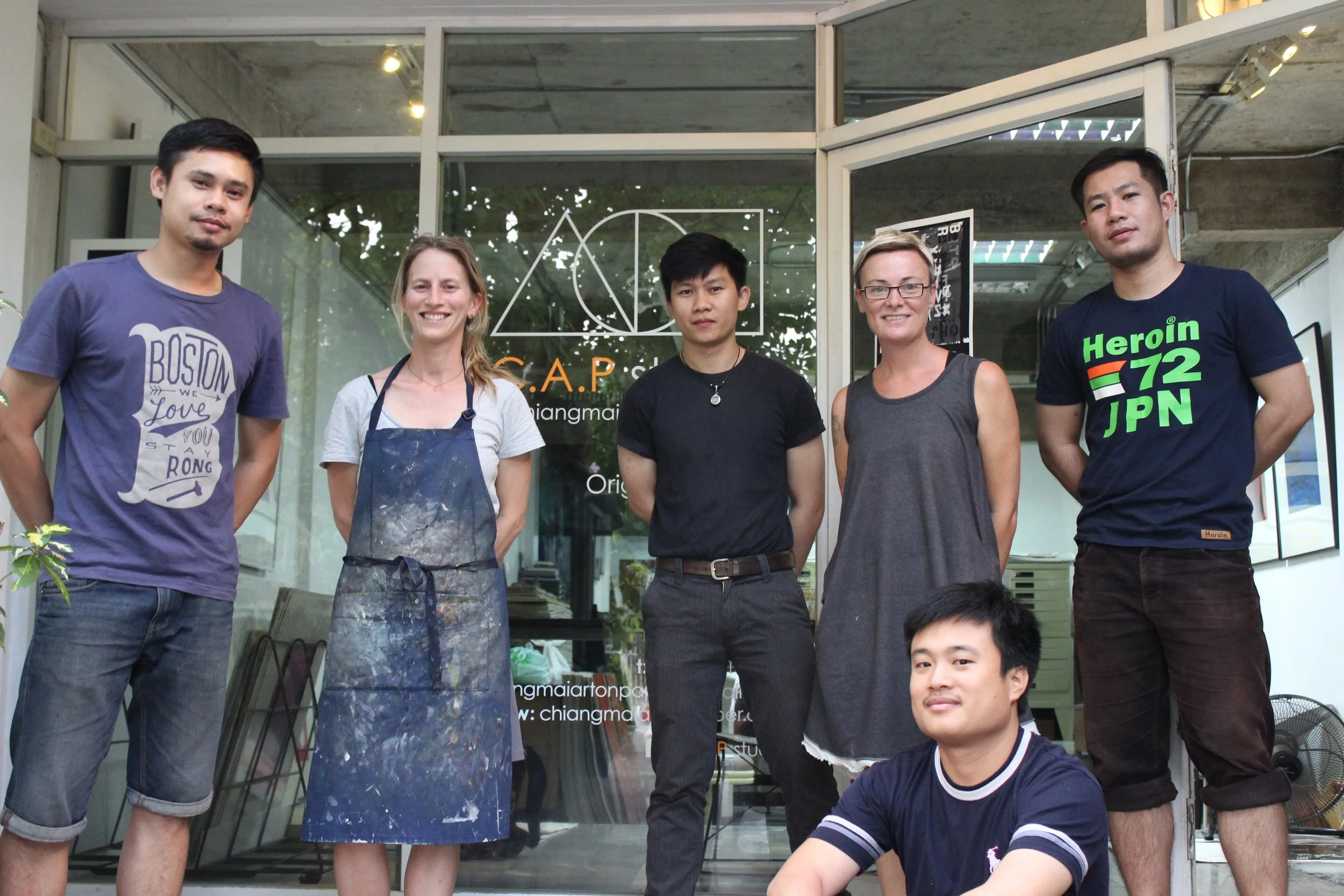 Group of six artists standing and sitting in front of an art studio, with a glass door behind them showing a logo for C.A.P. and some artwork inside.