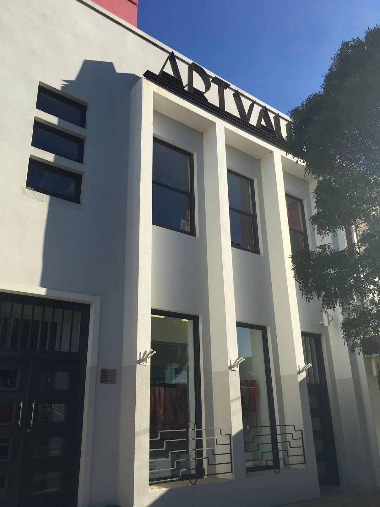 White building with large windows, a sign on top with partially visible black letters, and a tree on the right side.