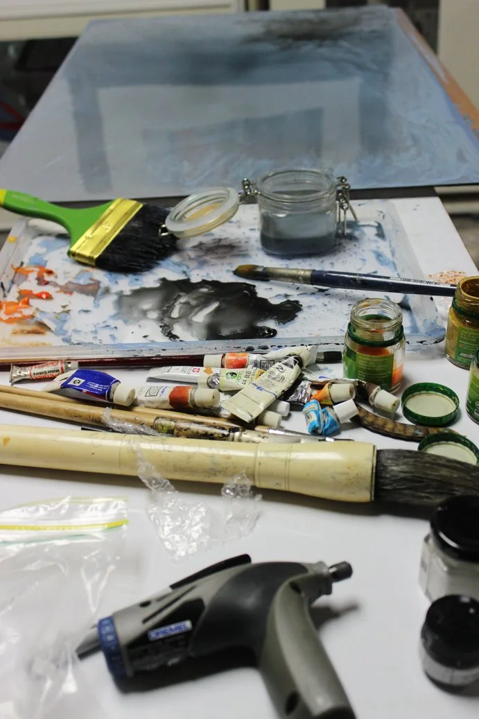 Art supplies including paintbrushes, acrylic paint tubes, jars of paint or ink, a palette, a paint roller, and a glue gun, with a canvas in the background.