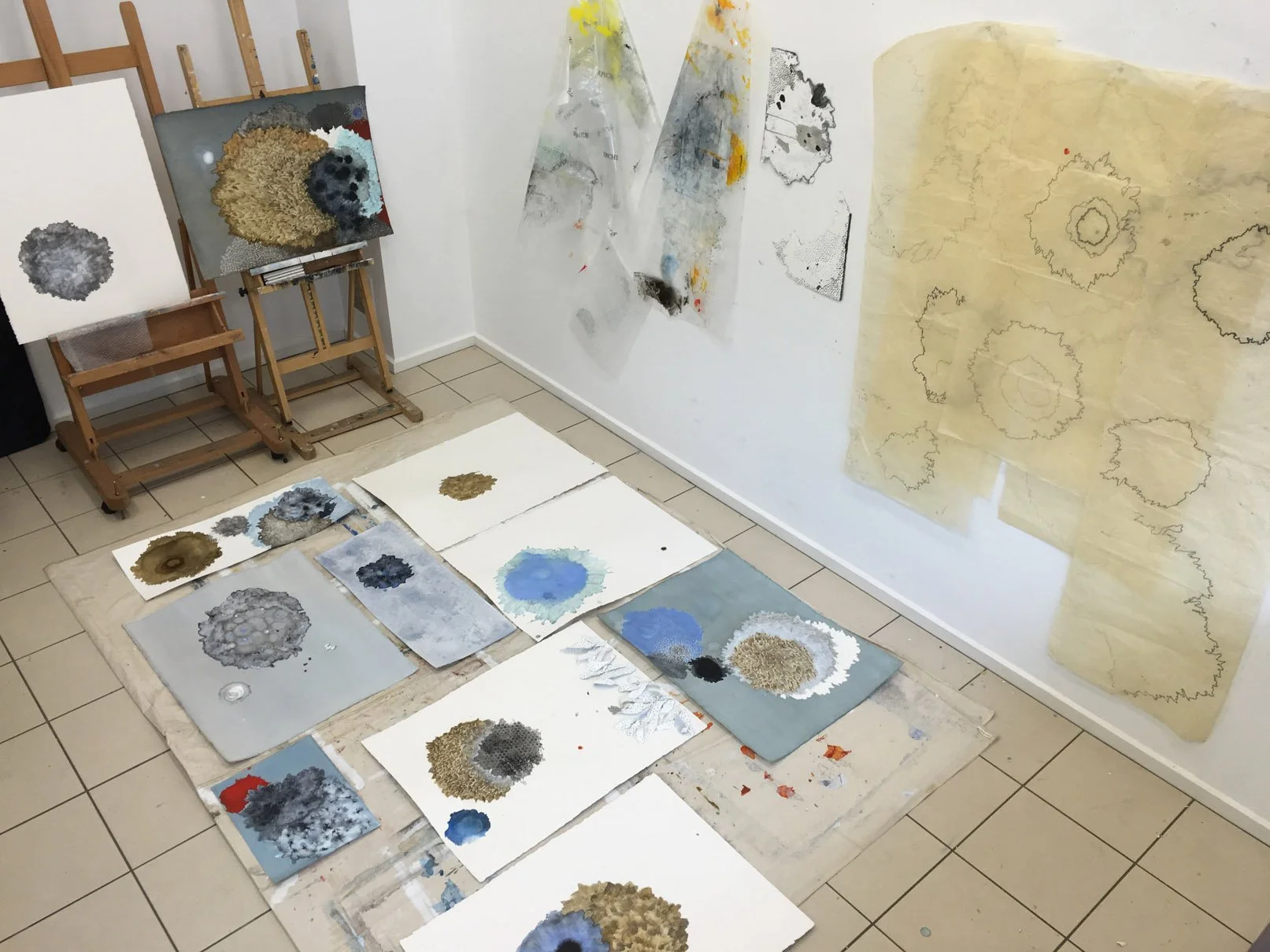 Art studio with multiple watercolor paintings on the floor and a few on the wall, featuring abstract cloud-like shapes in blue, gray, black, and tan colors. Several pieces are drying or in progress, and some are taped onto the walls.