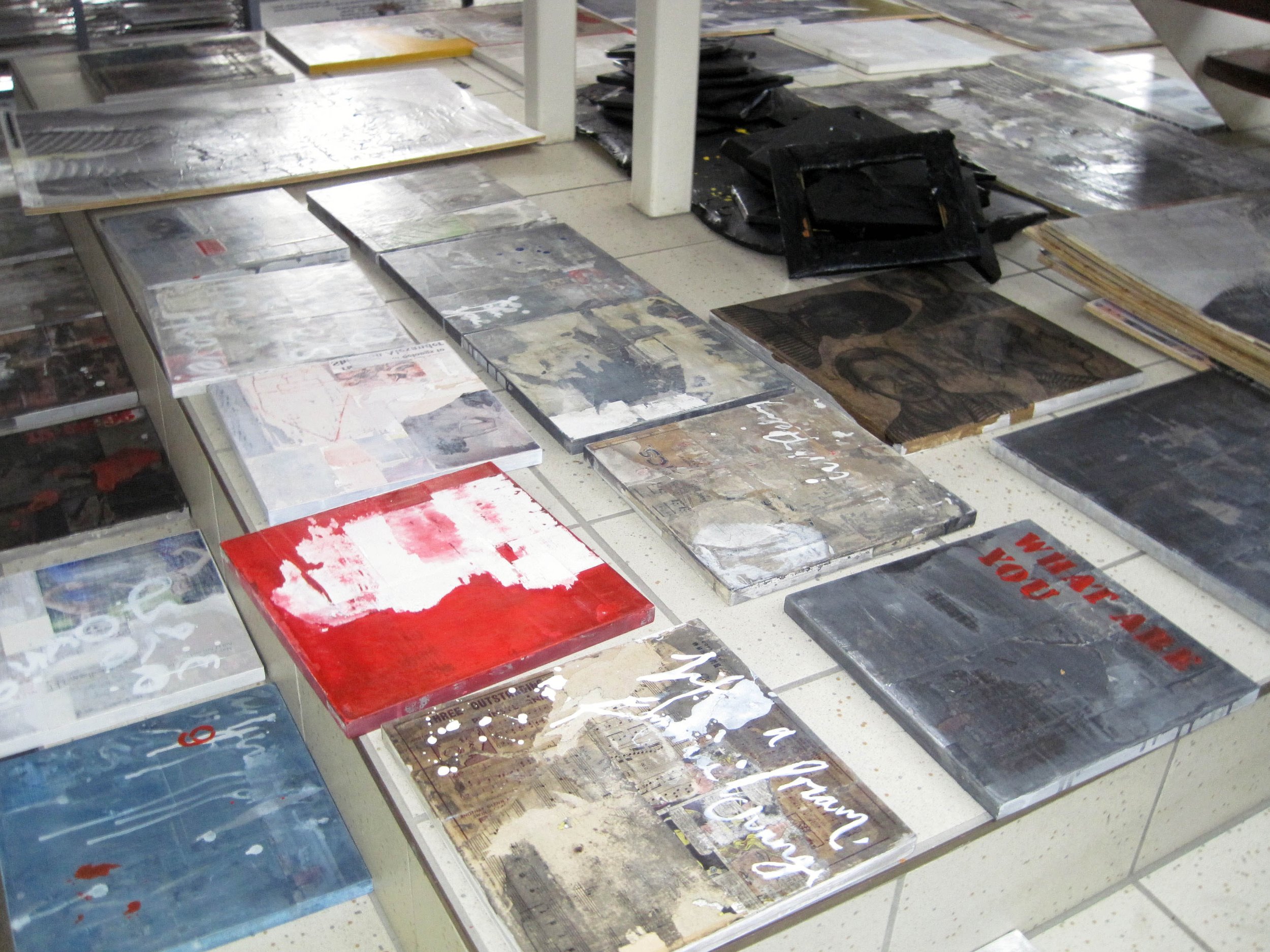 Art studio with multiple watercolor paintings on the floor and a few on the wall, featuring abstract cloud-like shapes in blue, gray, black, and tan colors. Several pieces are drying or in progress, and some are taped onto the walls.