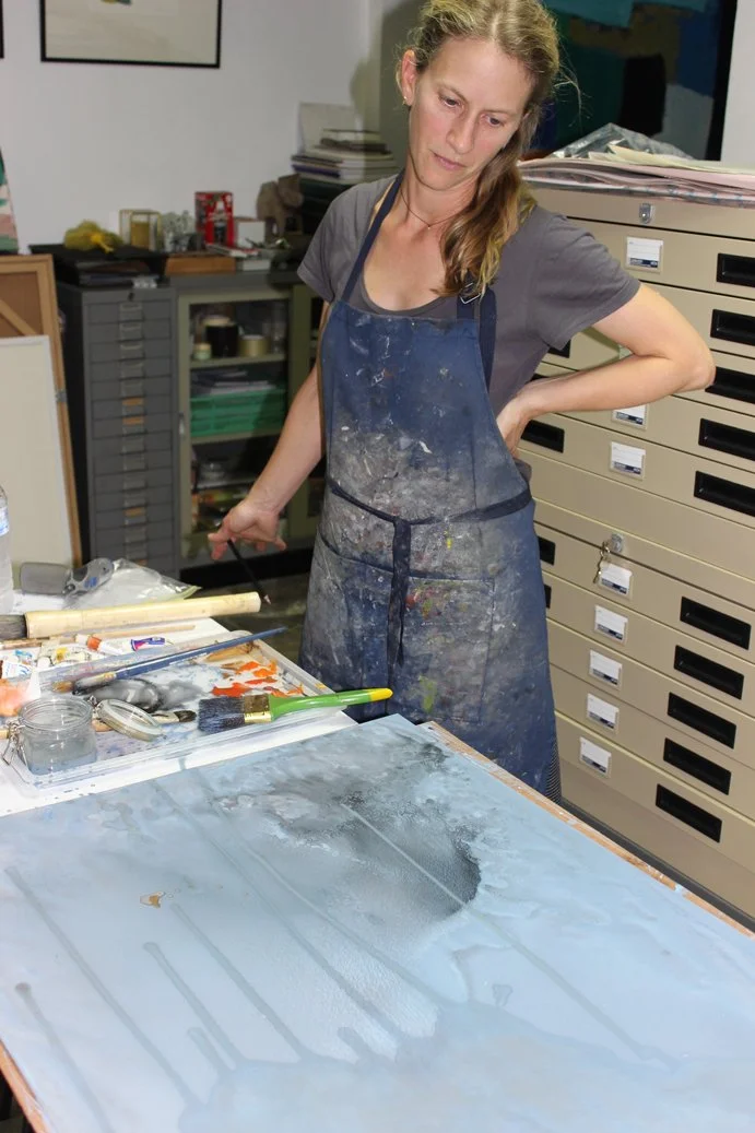 A woman wearing a paint-splattered apron looks down at her worktable in an art studio.