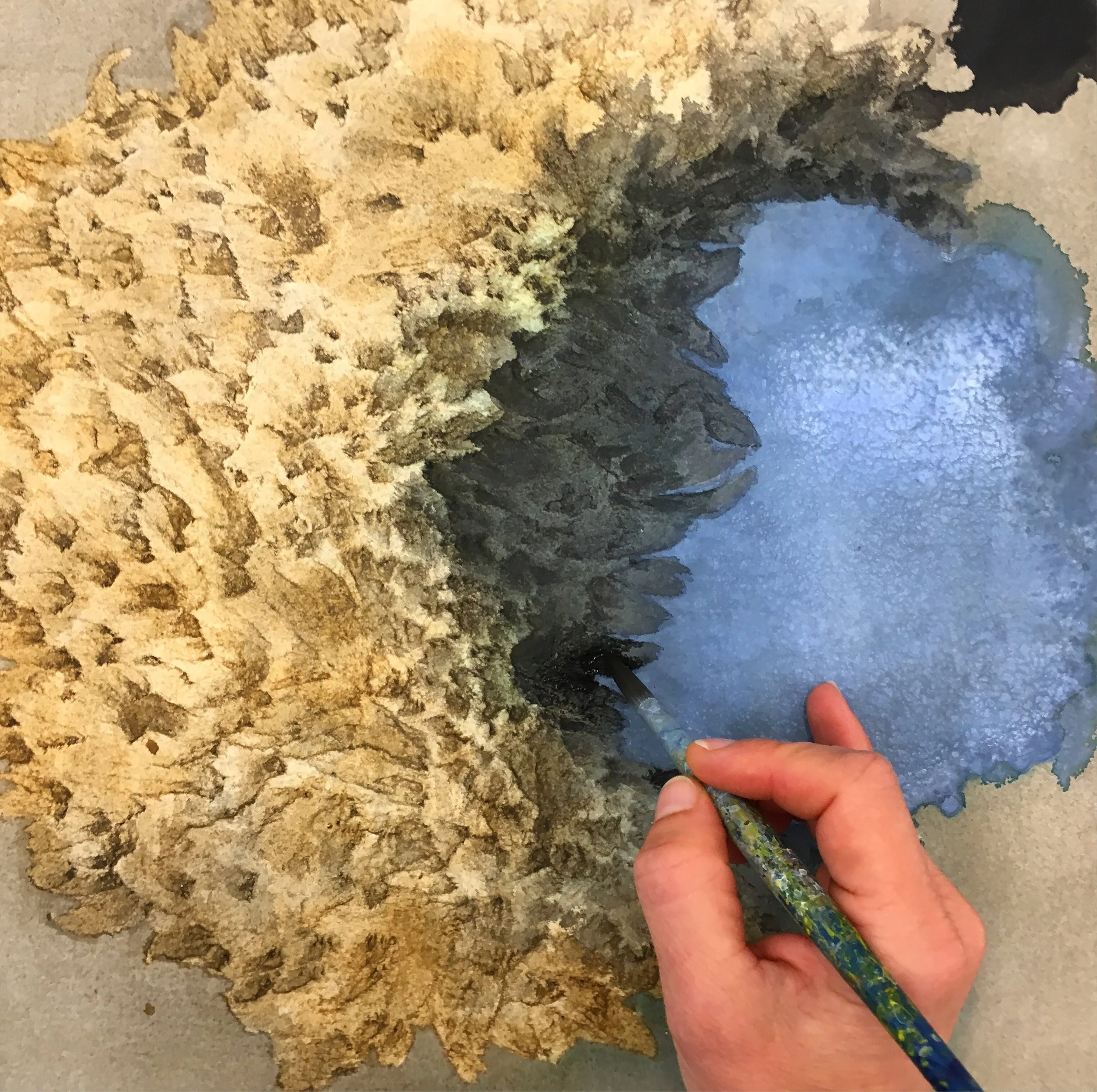 A person is painting a landscape scene featuring a rocky shoreline, a body of water, and a cloudy sky. The artist is holding a paintbrush and adding details to the water area on the canvas.