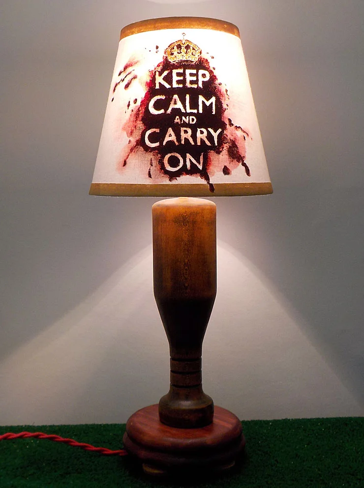 Hand painted shade with the "Keep Calm and Carry On" motivational saying, first printed on posters by the British government in preparation for the Second World War, on a red/blood splattered background. The base is made of Poplar wood turned on the lathe to look like a baseball bat and stained with amber brown and cinnamon red to make an antique finish.