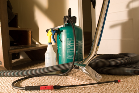 Carpet cleaning equipment including a professional vacuum, clear stain remover spray bottle and a green pump with spray nozzle.