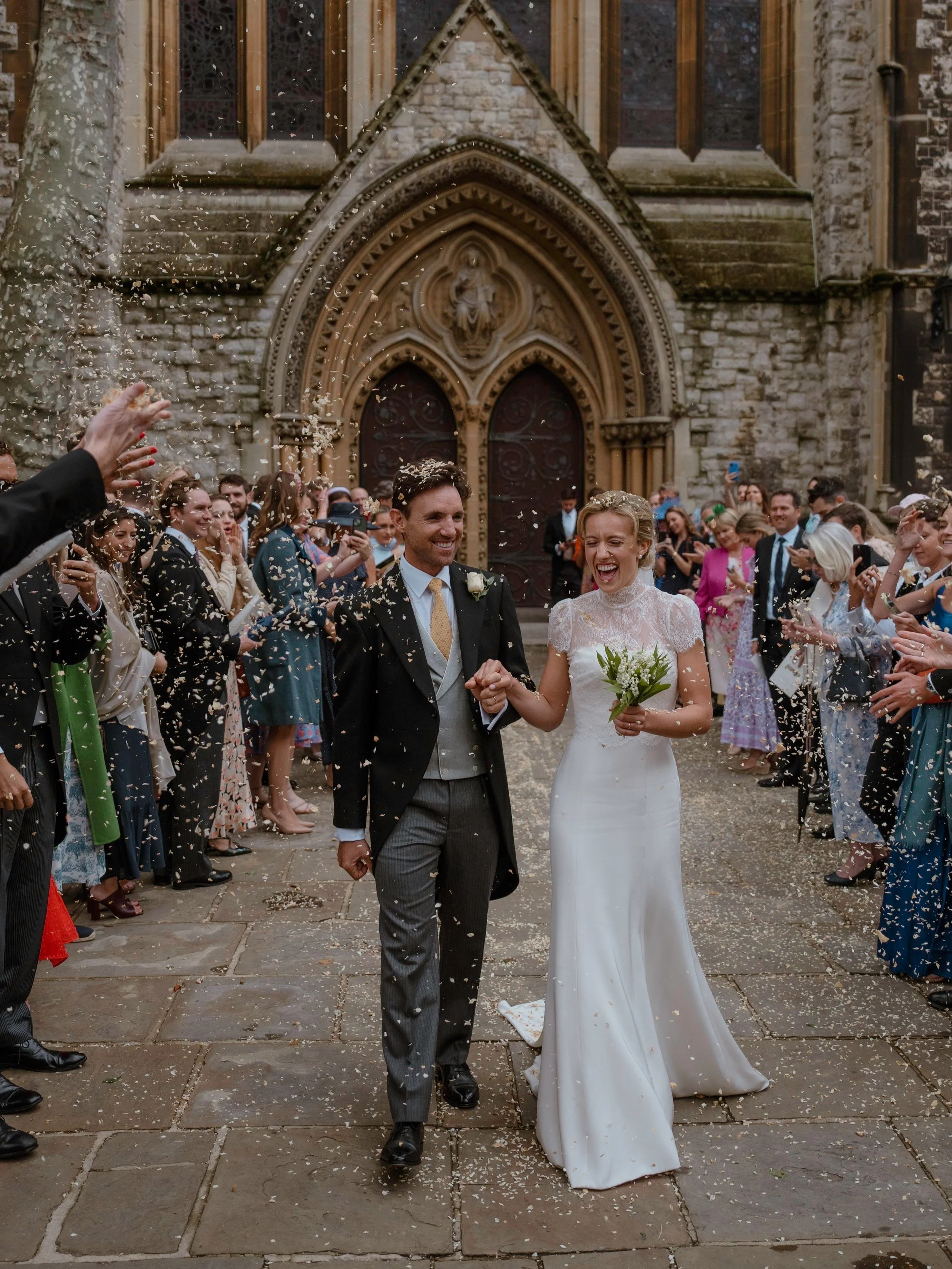Bride and groom walking hand in hand through a confetti tunnel outside a London church, laughing as guests celebrate around them. Documentary style wedding photography capturing natural emotion, movement and atmosphere in a classic London setting.