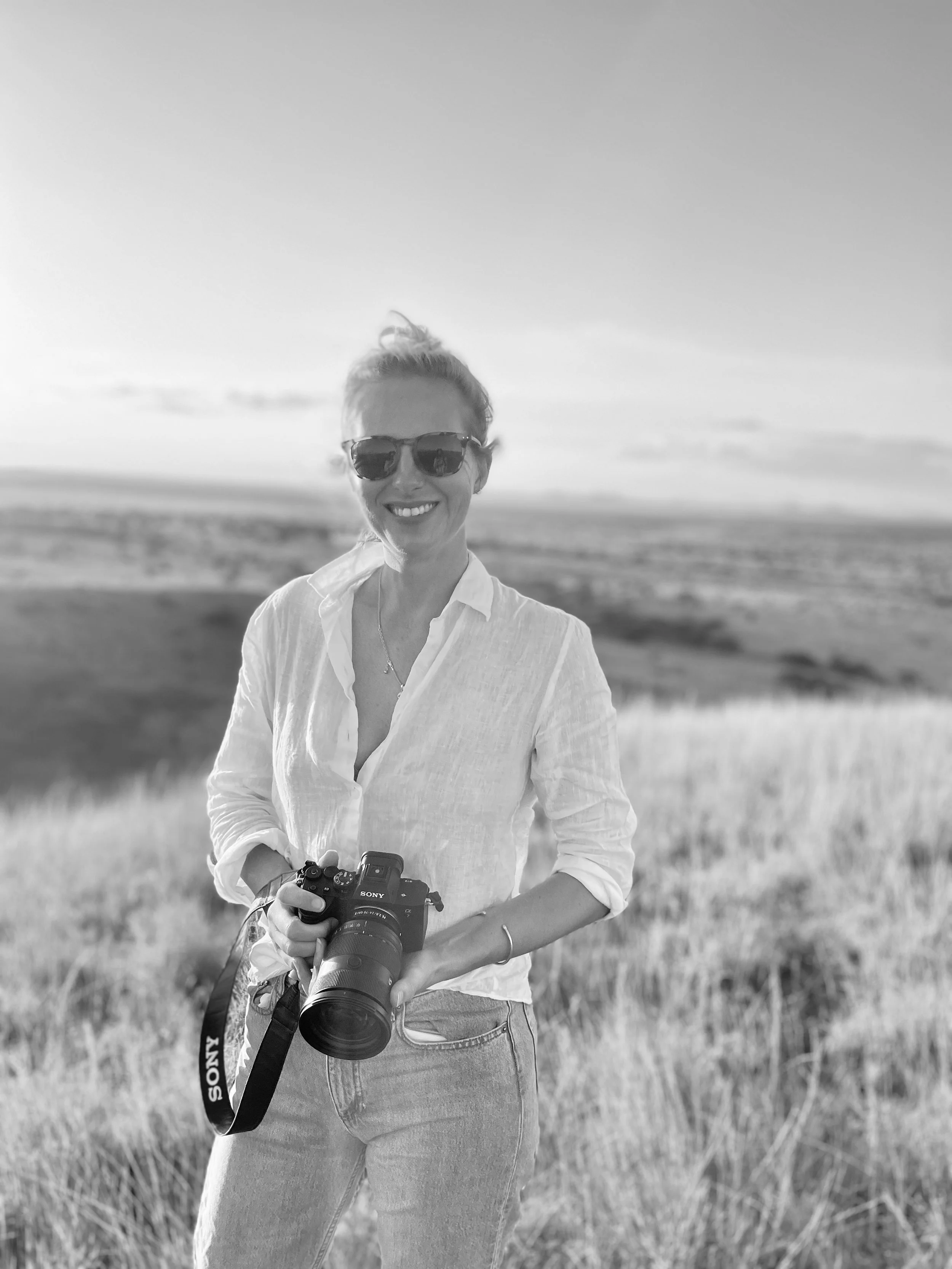 A woman standing outdoors in a grassy field holding a camera, wearing sunglasses and a light-colored shirt, smiling with the landscape in the background.