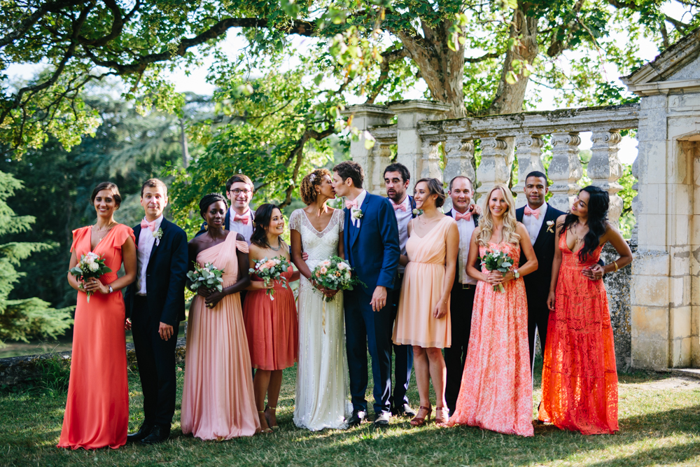 natural group photo of bridal party wearing orange and pink in front of trees and stone archway summer day