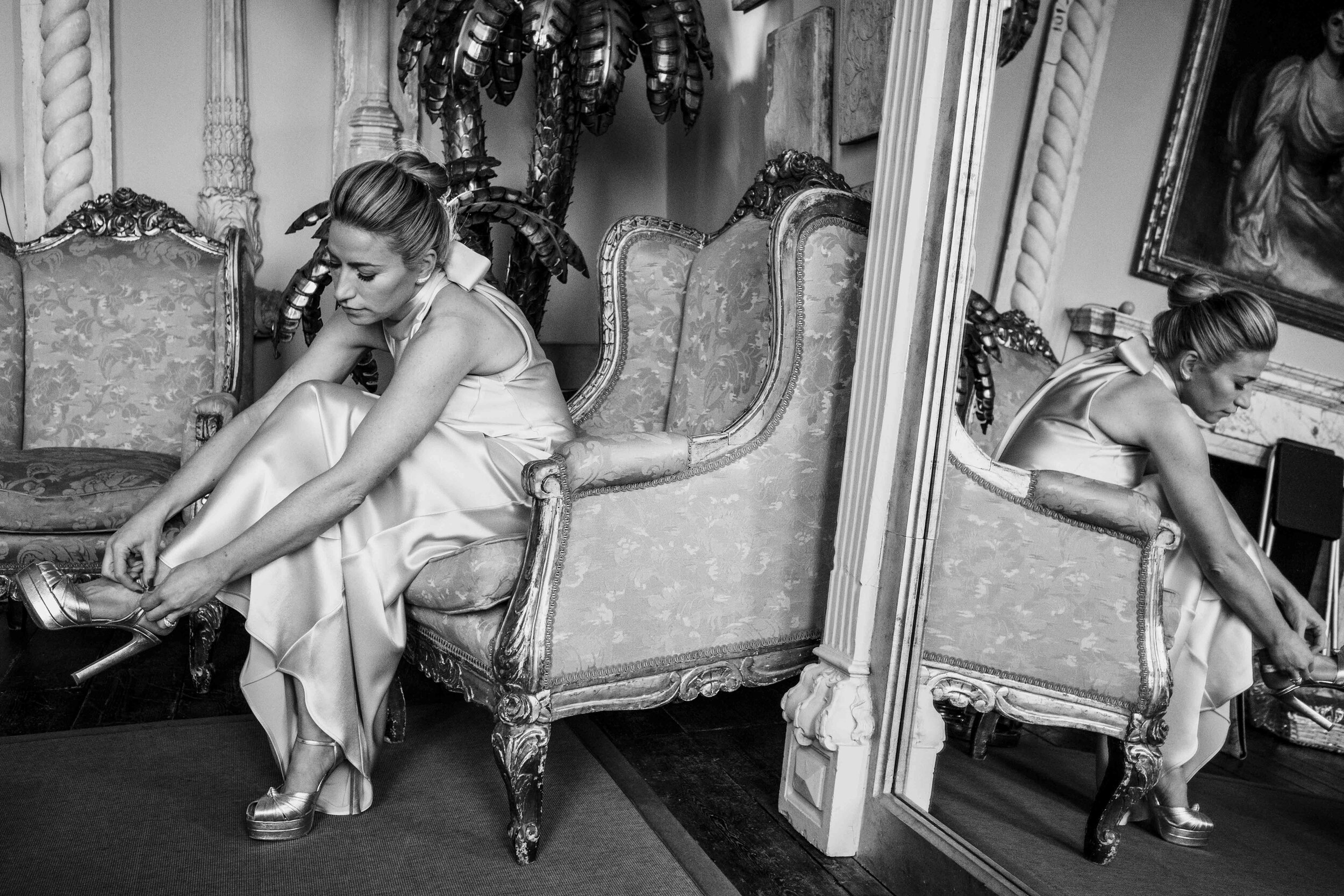 black and white picture of bride sitting in a chair doing up here shoe next to a mirror