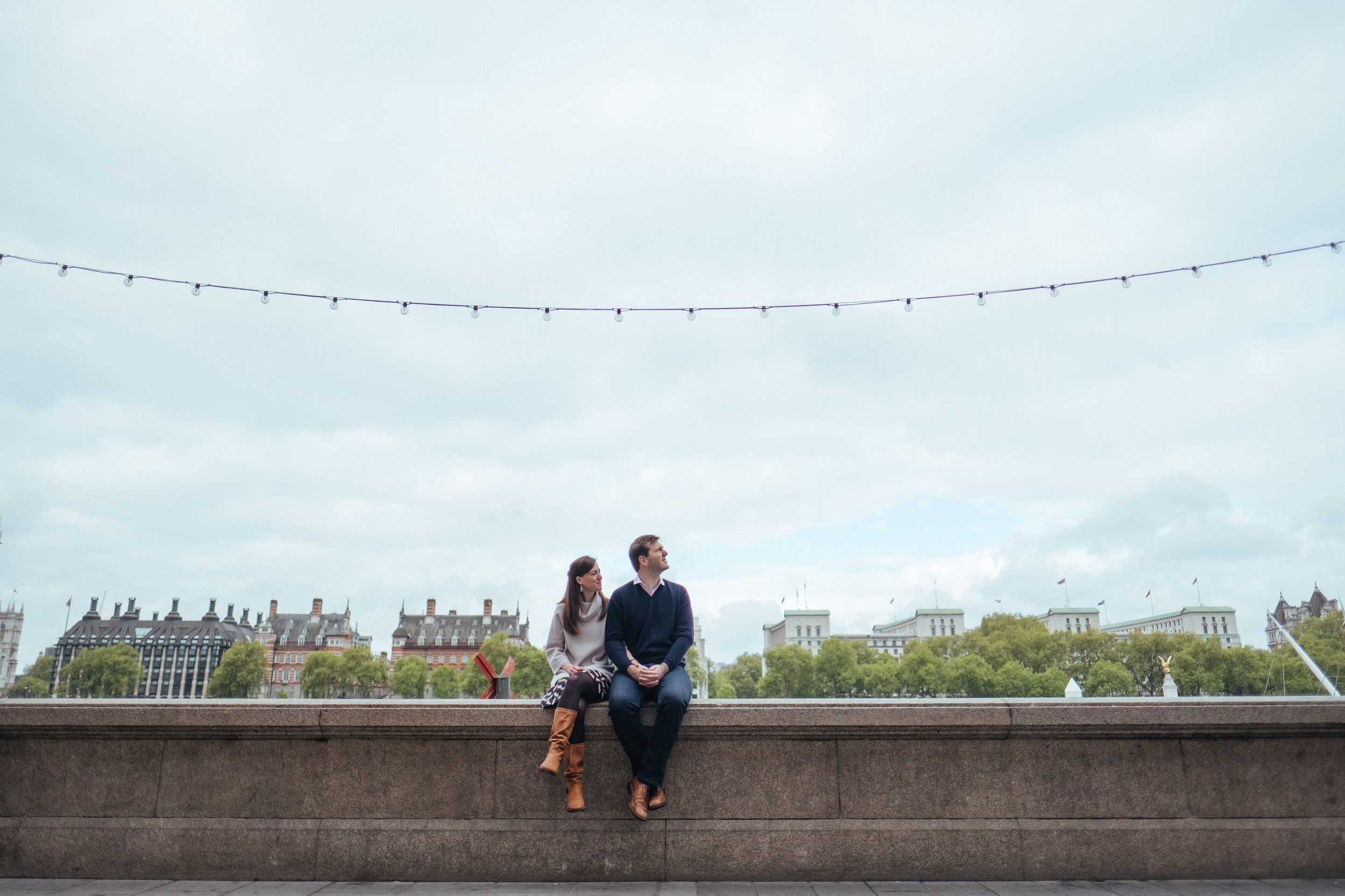 small couple sitting on edge of river with lots of sky and trees behind