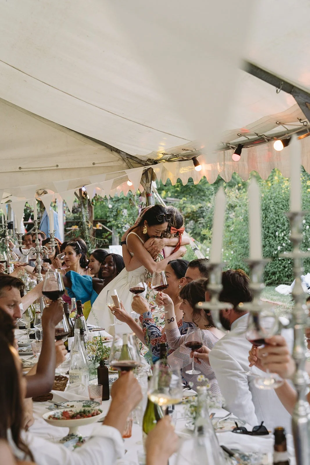 Bride leaning across a long table to hug a guest during the wedding breakfast while guests raise glasses around them.