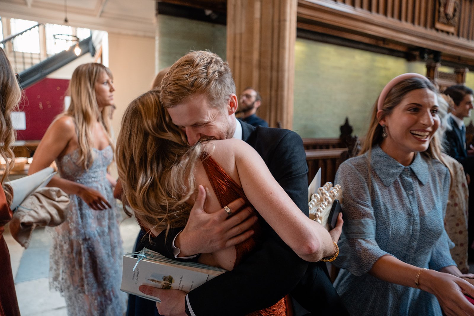 Wedding guests hugging and greeting each other inside a ceremony venue before the service.