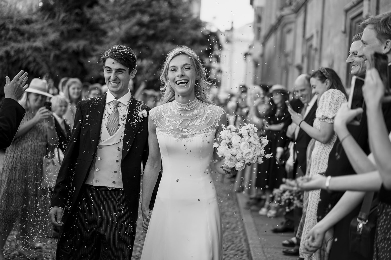Black and white photograph of a couple walking hand in hand through confetti outside a church surrounded by guests.