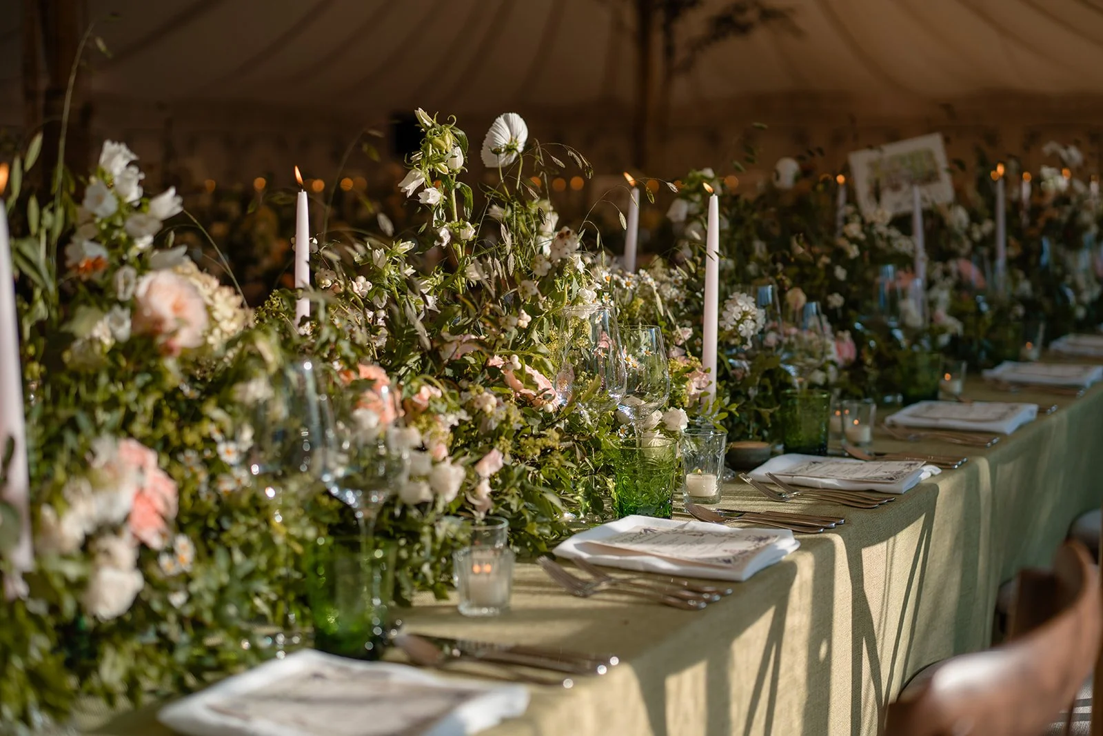 Long wedding reception table with green linen, floral arrangements, tapered candles, glassware and place settings inside a marquee.