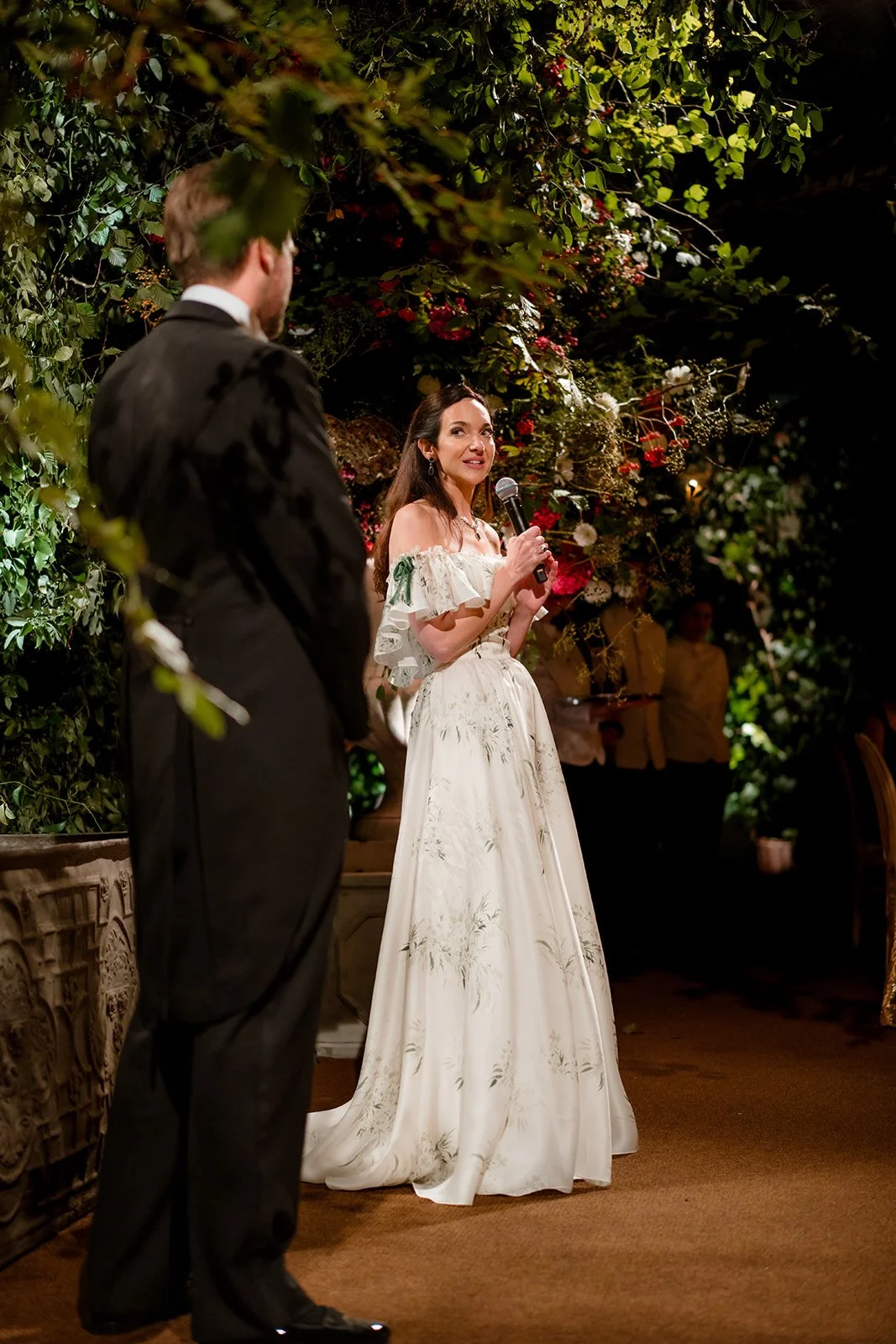 Bride delivering a speech during a candlelit wedding celebration surrounded by floral installations.