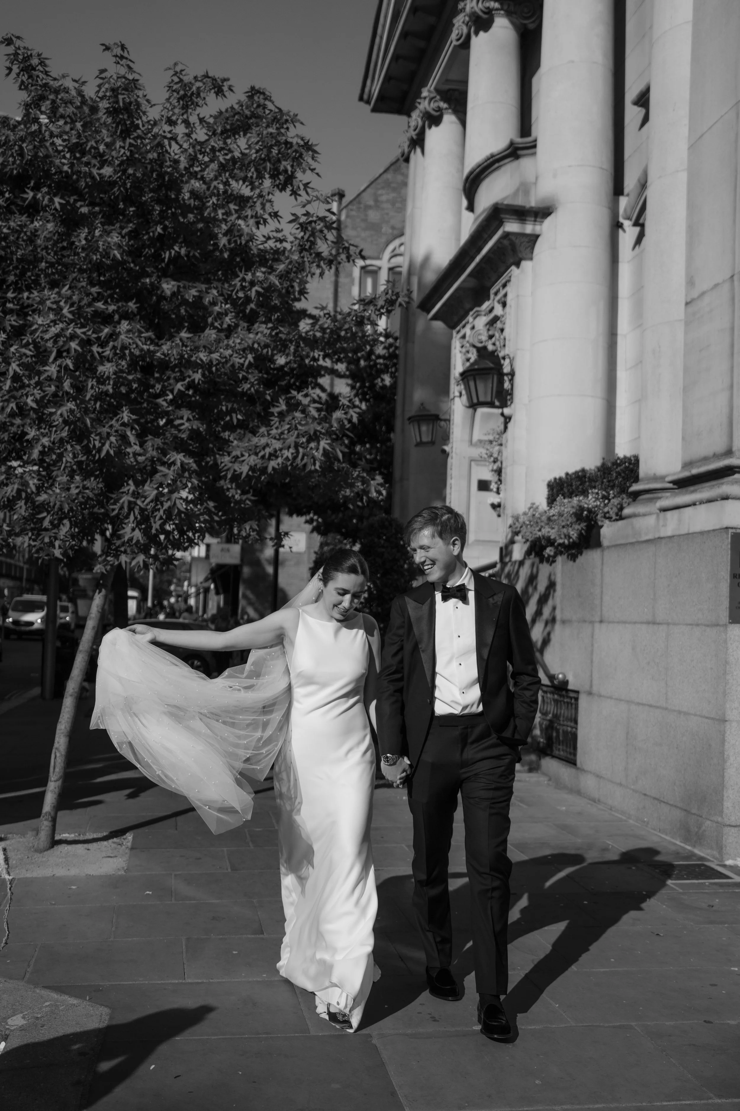 Bride and groom walking hand in hand along a London street after their wedding ceremony, the bride lifting her veil as sunlight creates long shadows. Black and white documentary style wedding photograph showing relaxed, joyful movement in the city.