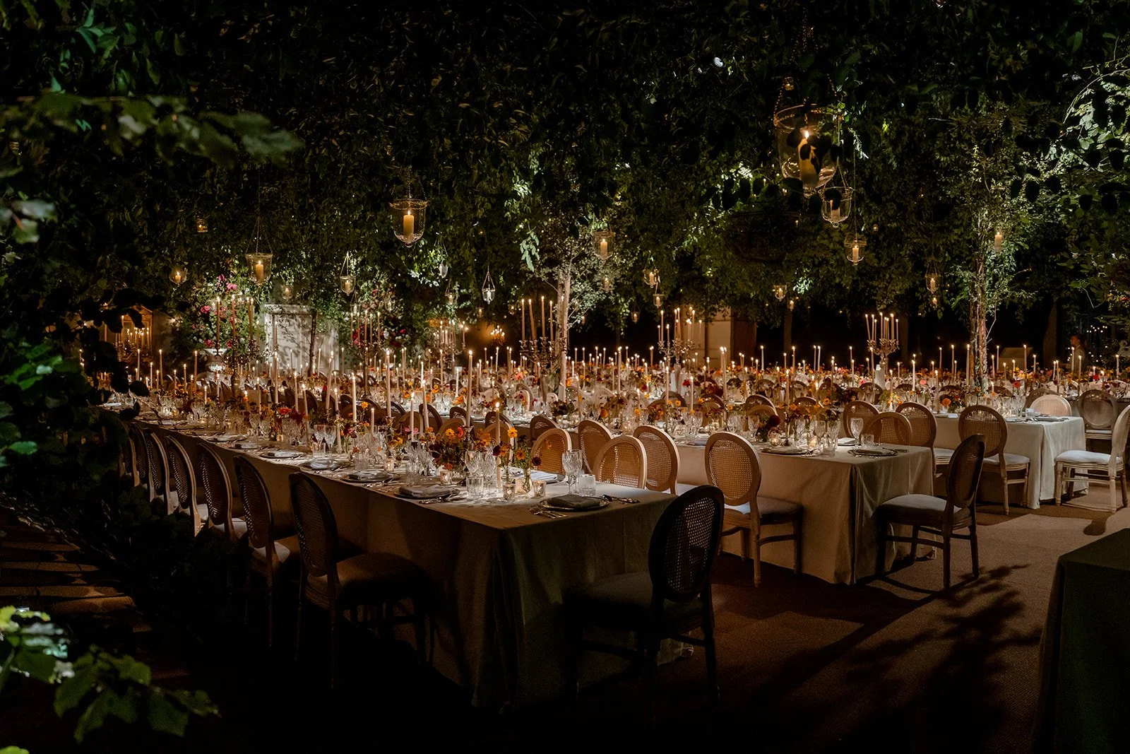 Long candlelit banquet tables set beneath hanging greenery for an elegant evening wedding reception.