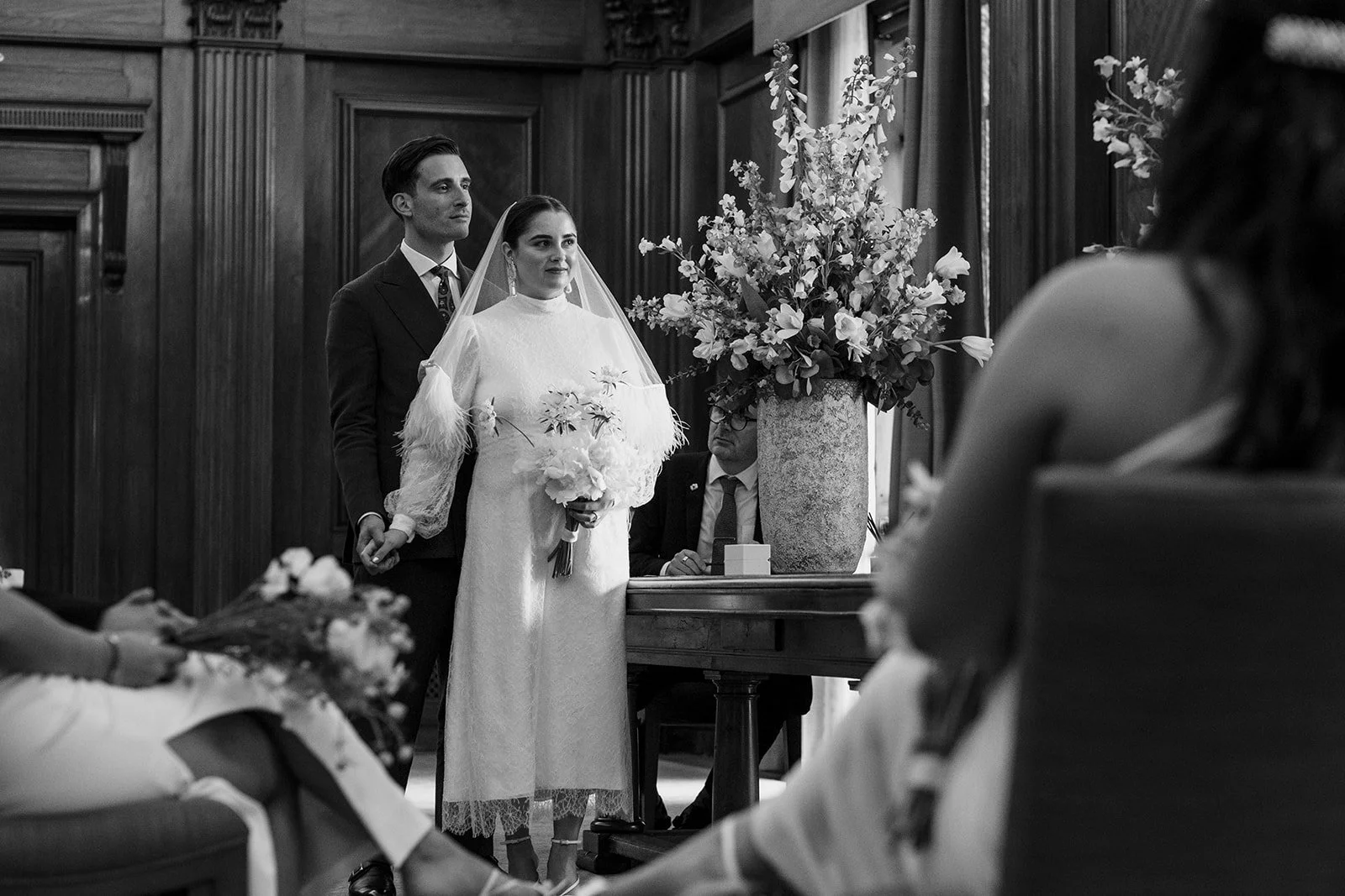 Couple standing during wedding ceremony in wood panelled London room with floral arrangement
