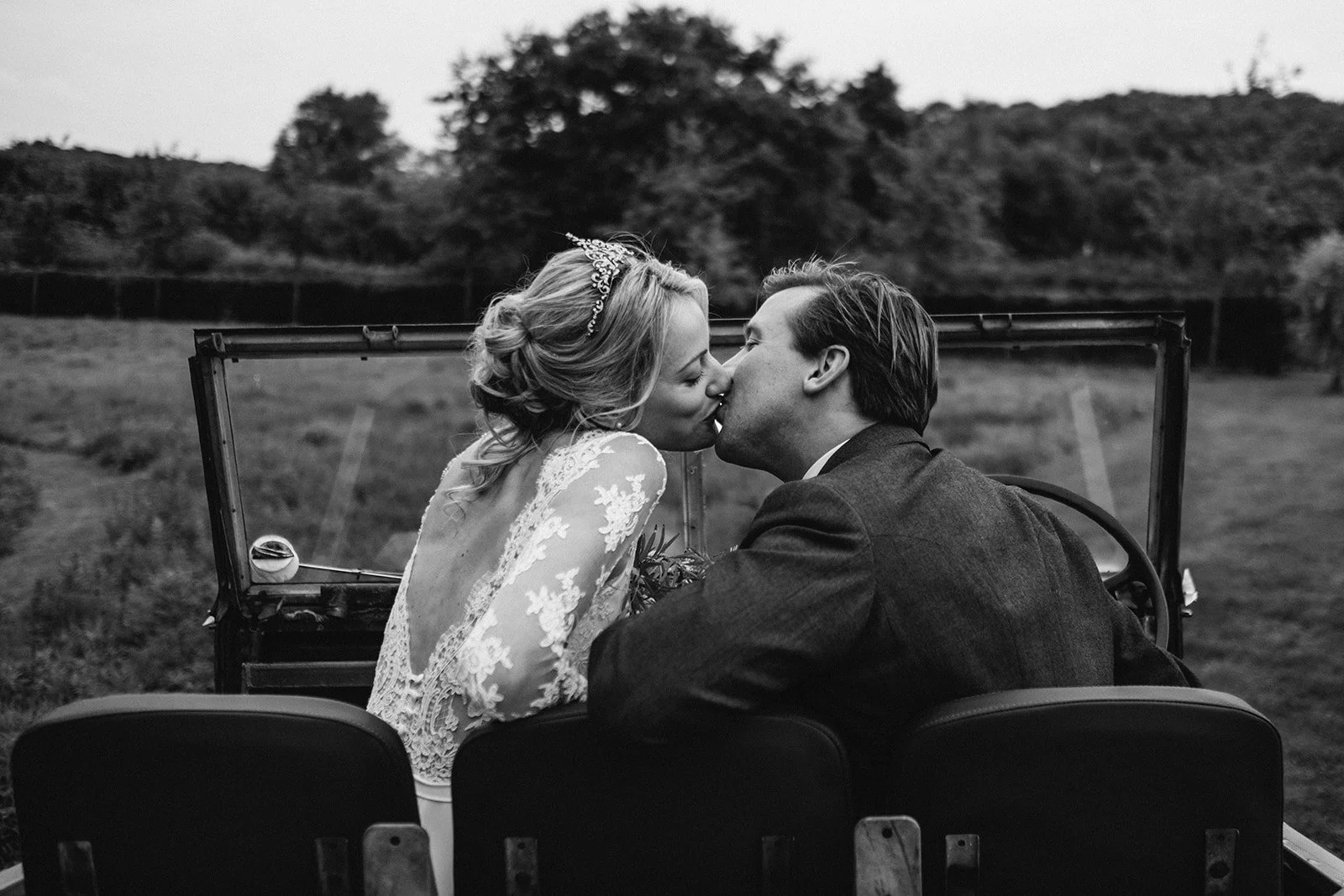Bride and groom kissing in the back of a vintage car in a field, photographed in a documentary wedding photography style
