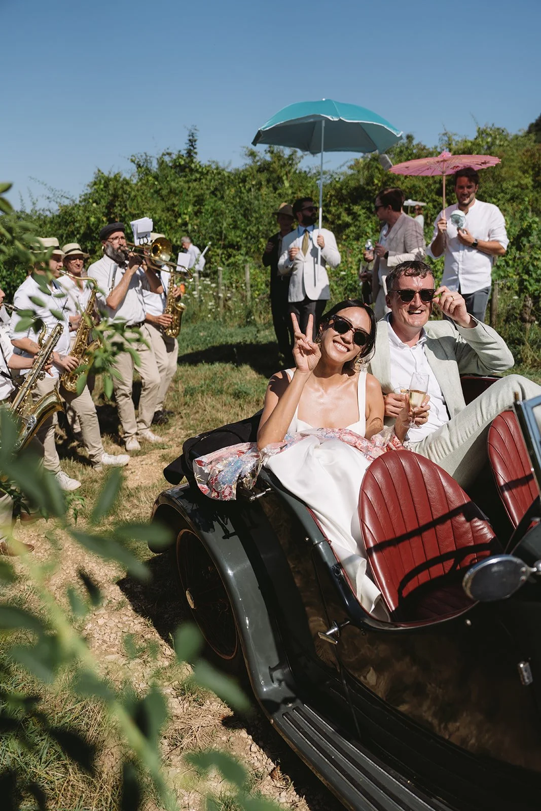 Bride and groom laughing in a vintage car as guests and musicians walk behind during an outdoor wedding procession