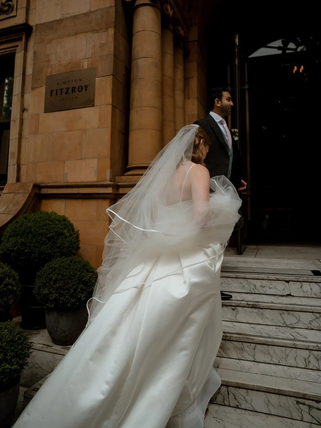 Bride walking up the marble steps of the Kimpton Fitzroy hotel in London, her veil and silk gown catching the light as she and her partner enter the venue. Candid documentary wedding photograph capturing movement and elegant city architecture.