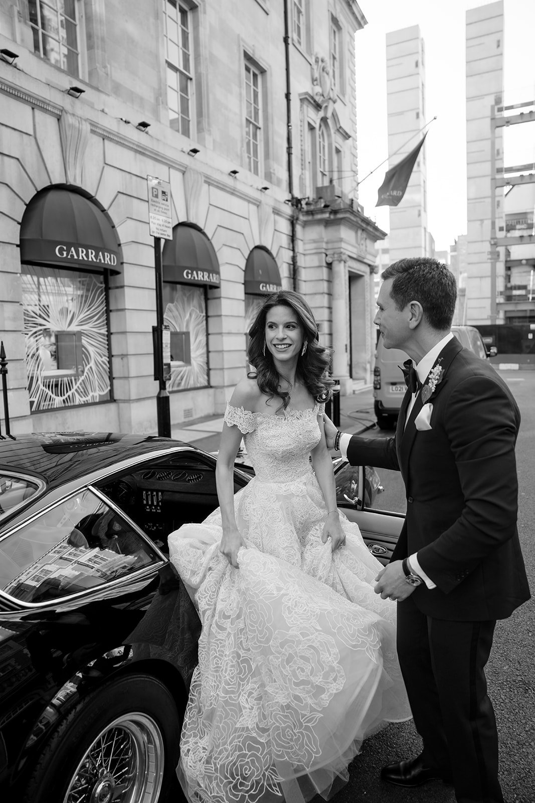 Bride and groom standing beside a classic car on a London street after their wedding ceremony.