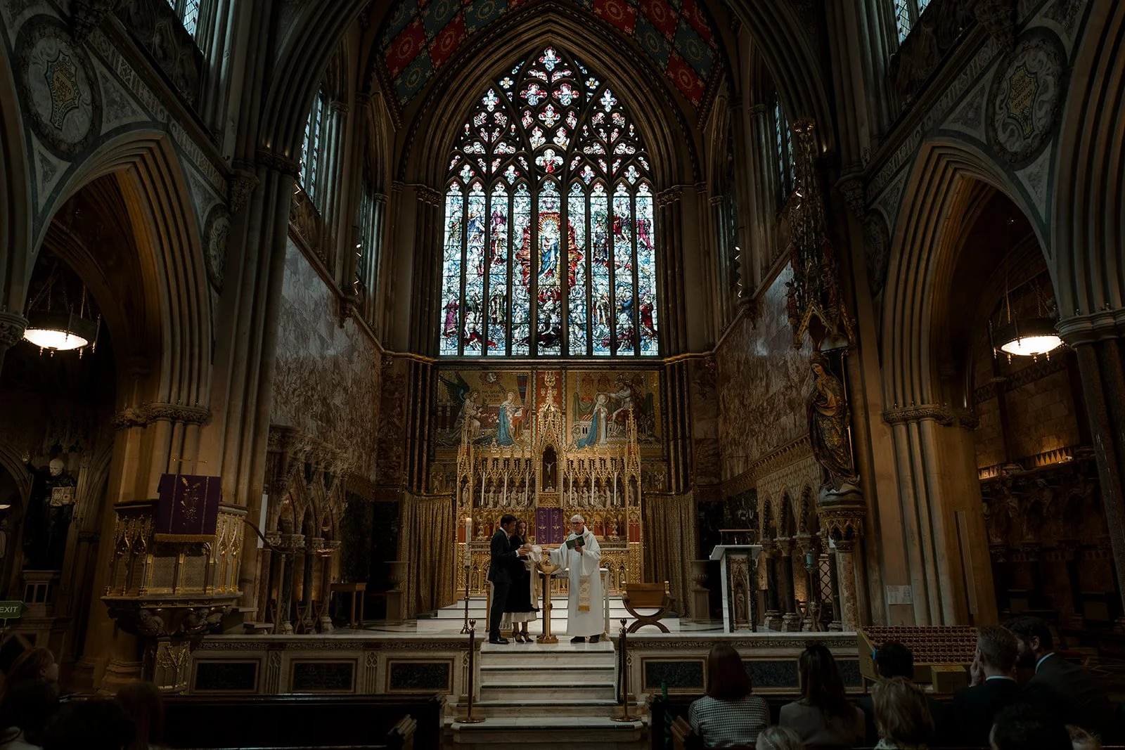 Christening ceremony taking place at the altar inside Farm Street Church in Mayfair, London, with stained glass windows and ornate Roman Catholic interior.