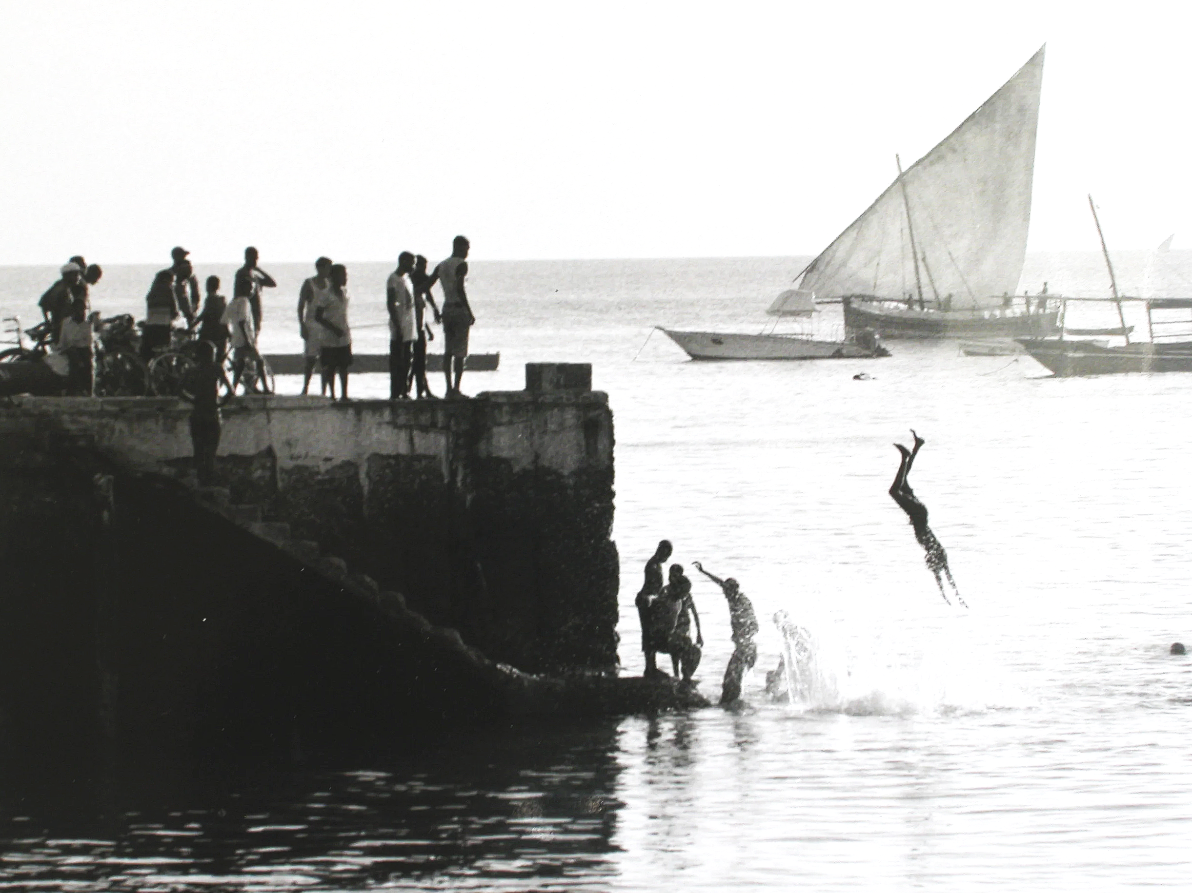 People standing on a pier near the water, some on bicycles, with boats on the water and a sailboat in the distance in Zanzibar, East Africa.