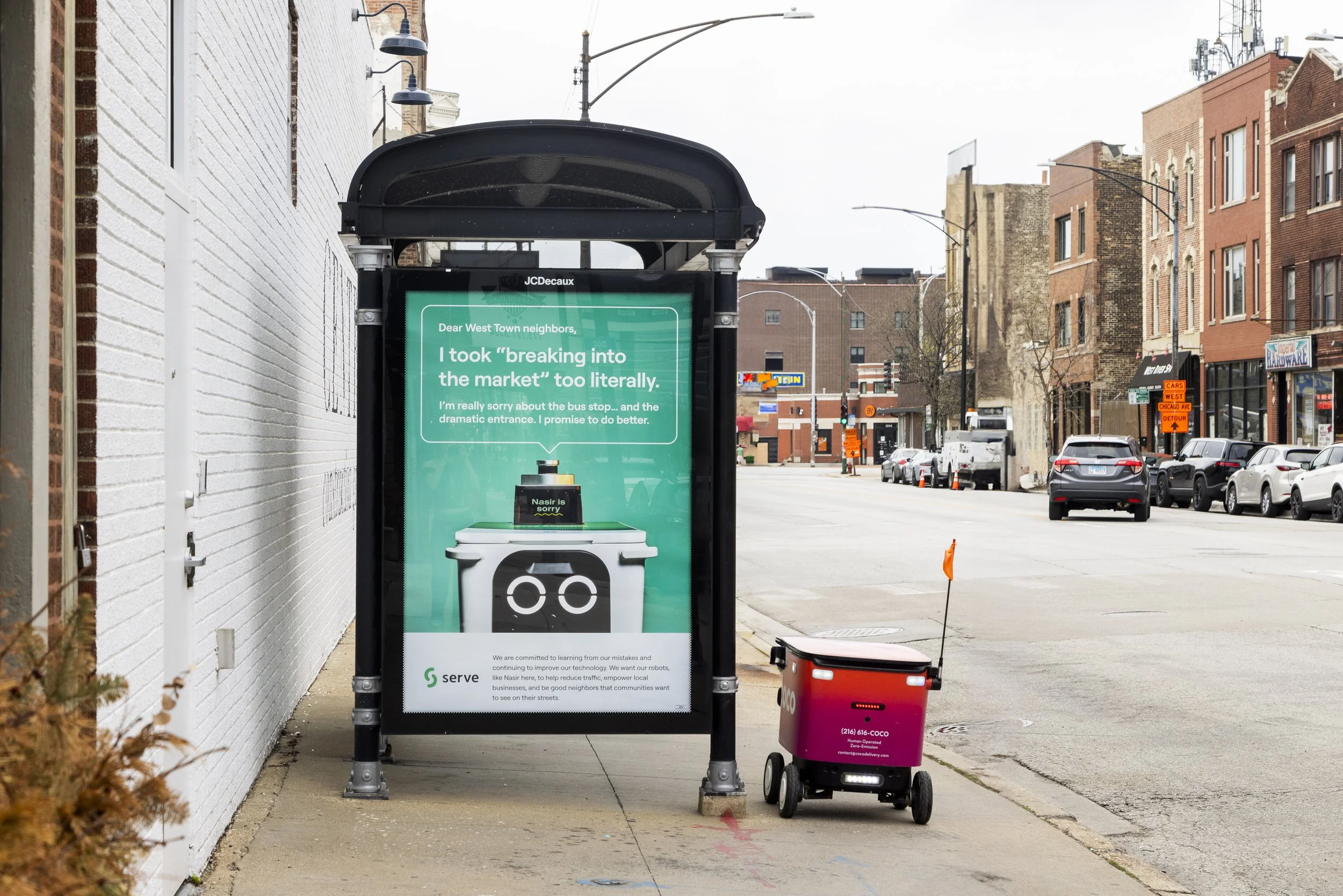 Bus stop shelter with a digital display screen showing a message to West Town neighbors, with a sidewalk, parked cars, and buildings in the background.