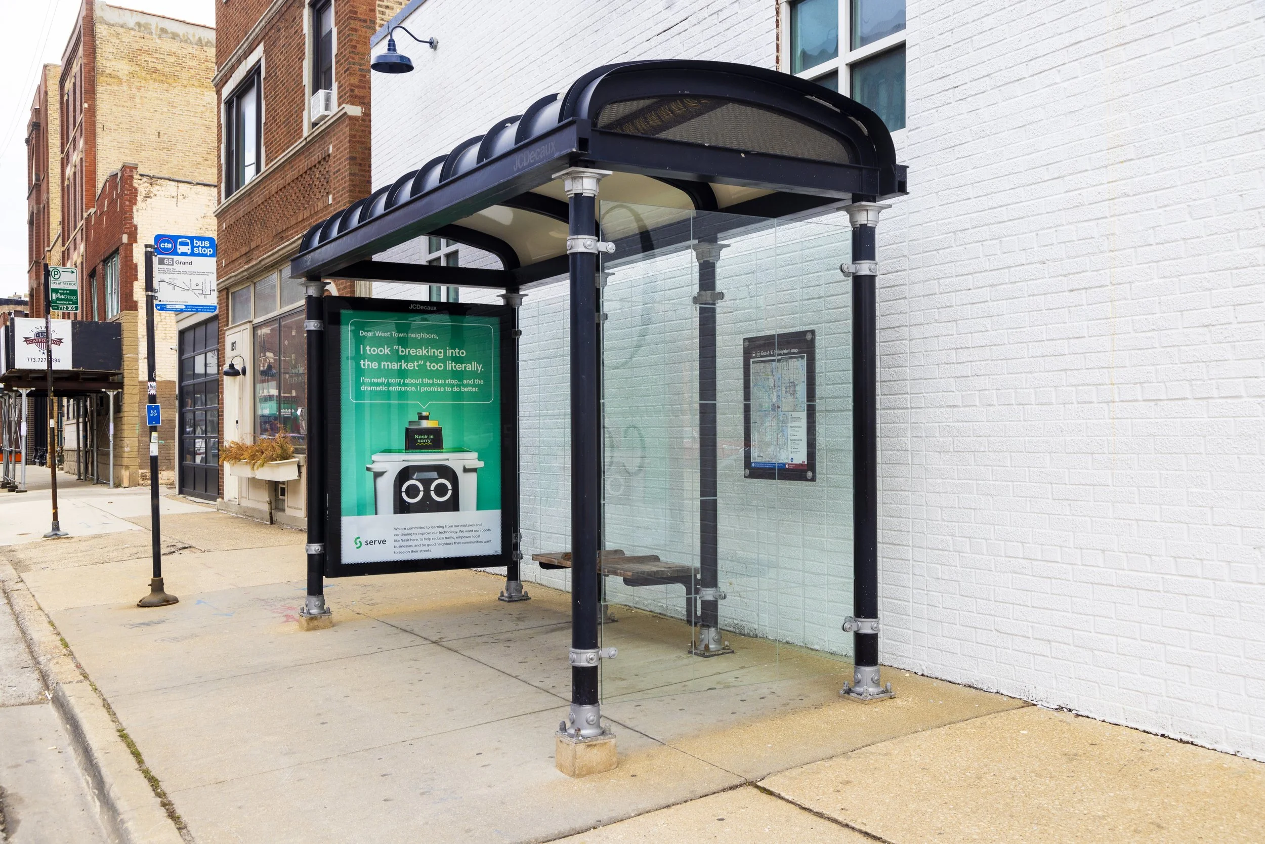 Bus shelter with transparent glass panels, a black rubber roof with a curved design, a digital advertising display, and a map inside the shelter, situated on a sidewalk along a brick building and a white painted wall.
