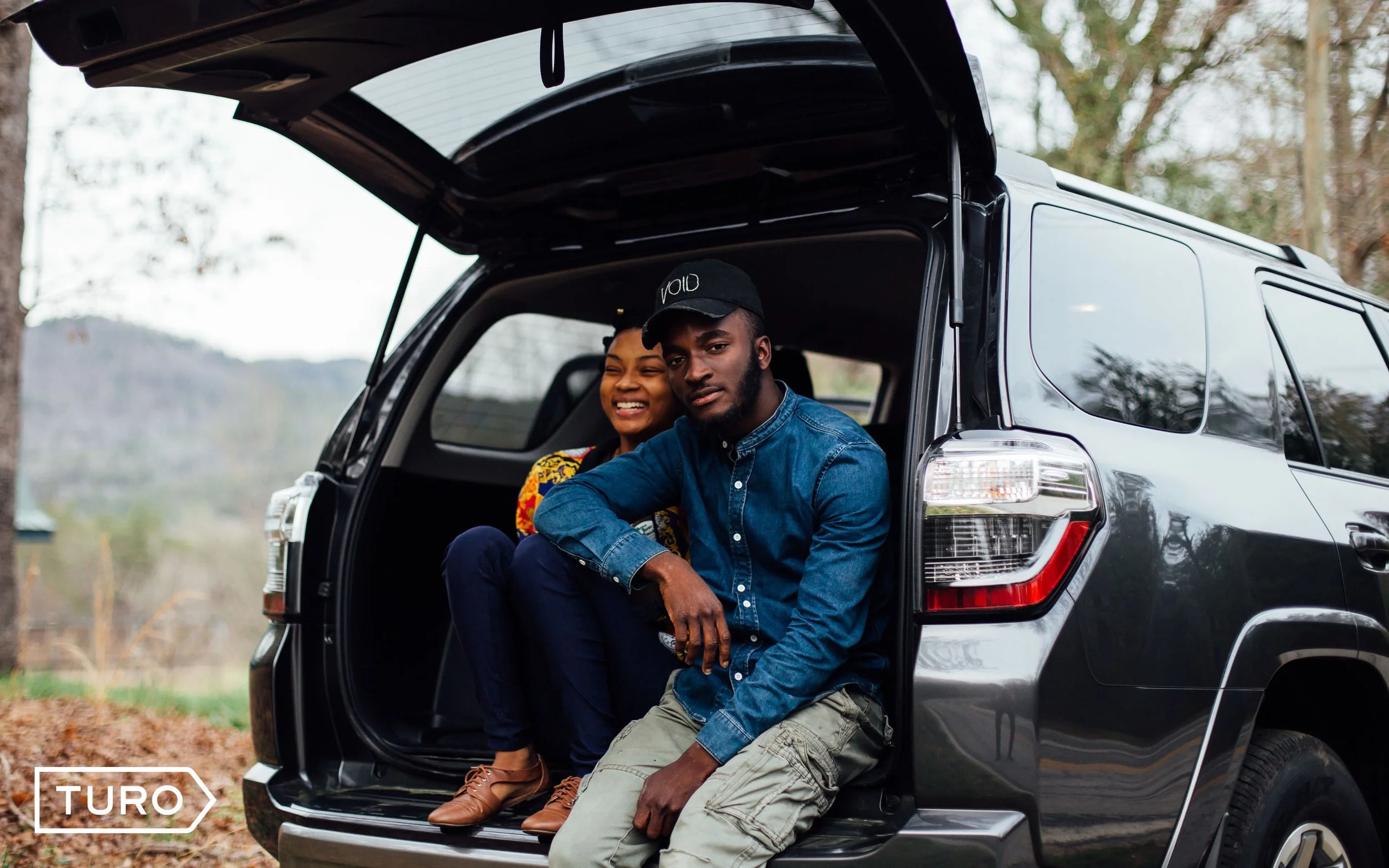 Two people sitting in the open trunk of a black SUV, with a background of trees and outdoor scenery.