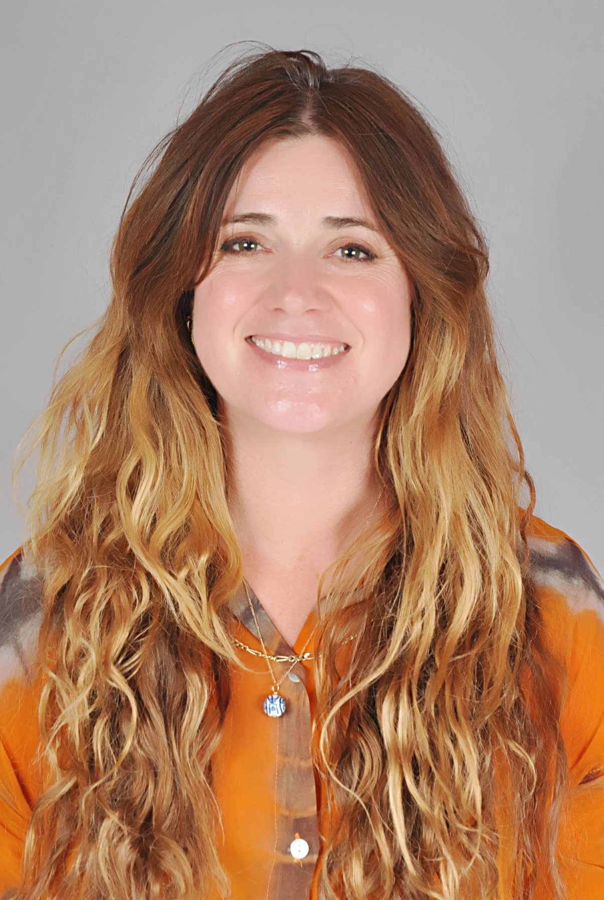 A woman with long, curly, reddish-blonde hair wearing an orange and brown blouse, smiling at the camera against a gray background.
