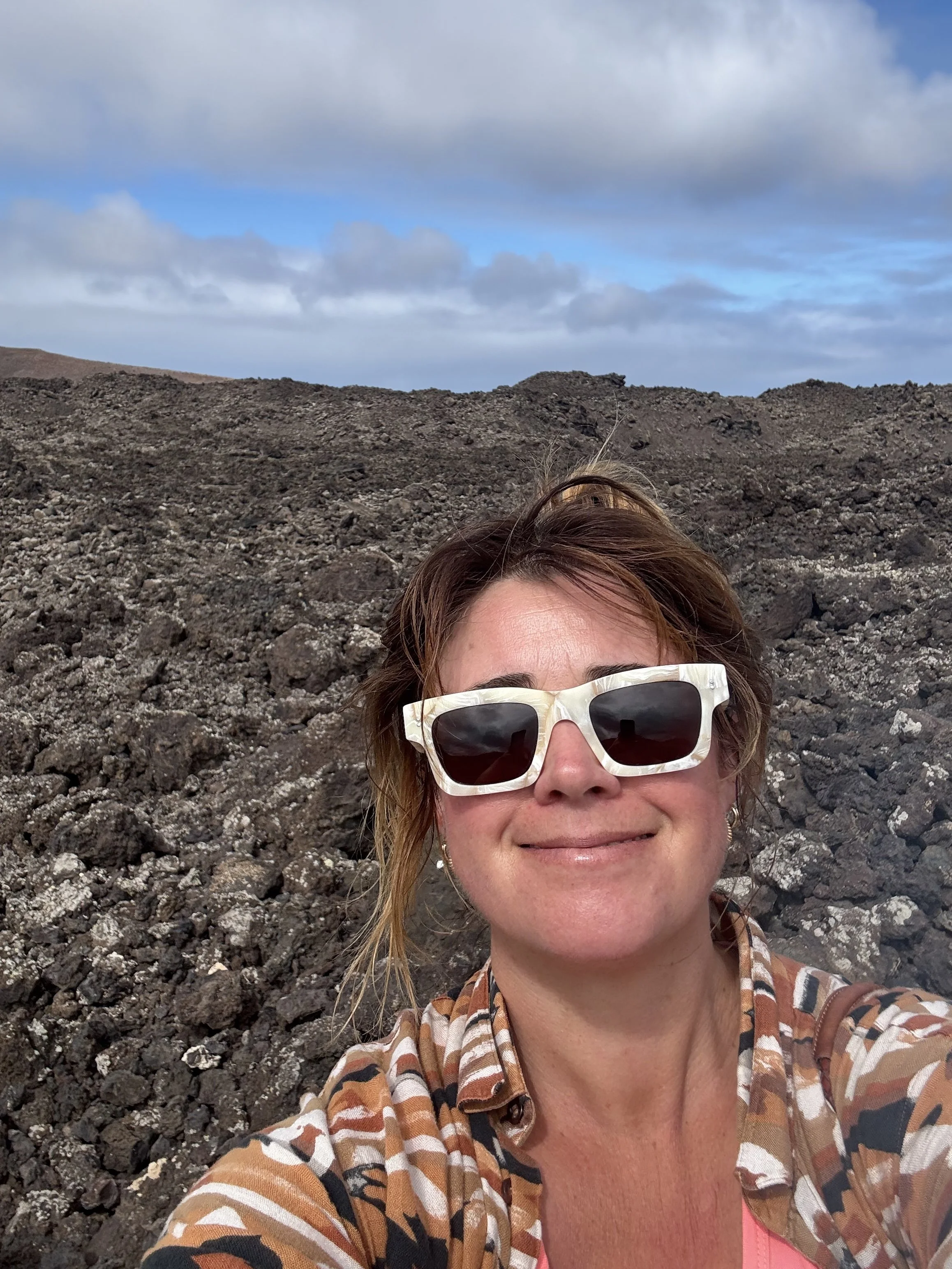 A woman with short brown hair wearing sunglasses, a striped brown and tan shirt, and a pink top, smiling while taking a selfie outdoors on a rocky volcanic landscape with a partly cloudy sky.