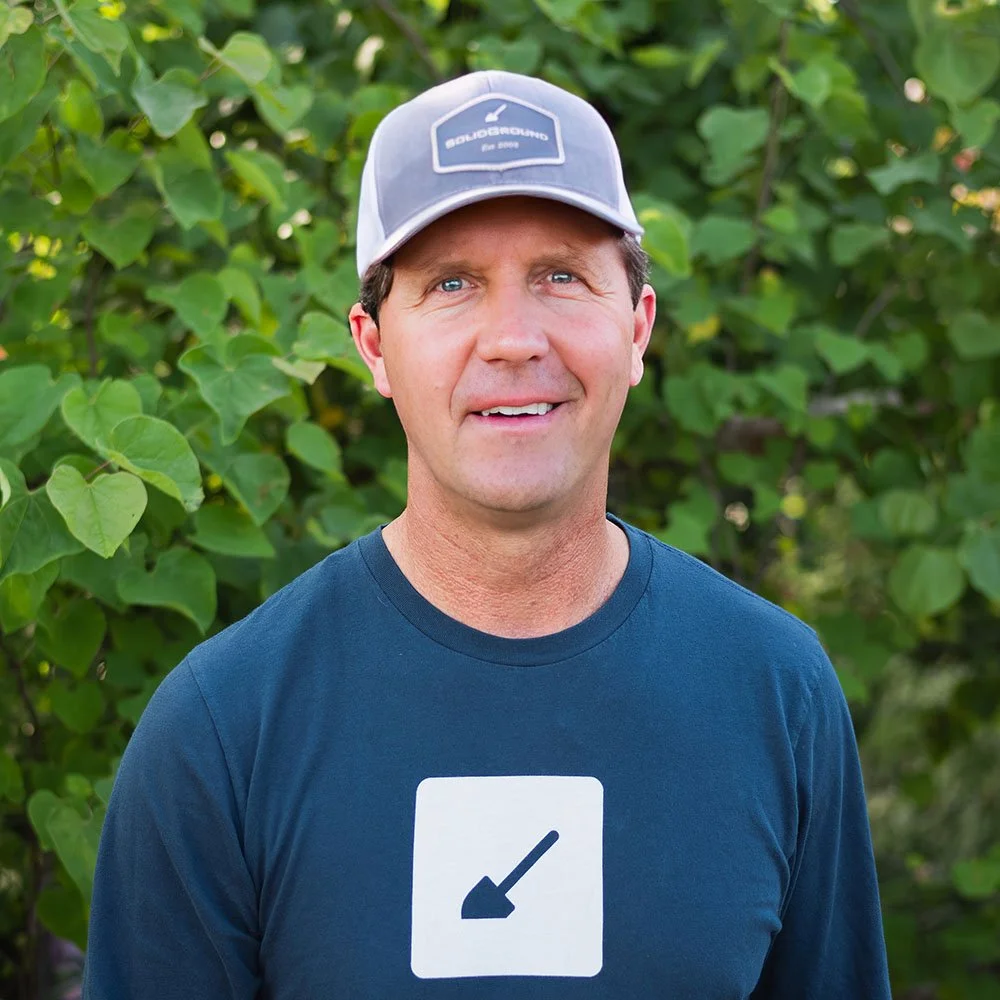 smiling caucasian male wearing gray cap with brim and blue shirt with white solid ground logo, standing infront of leafy green shrubs