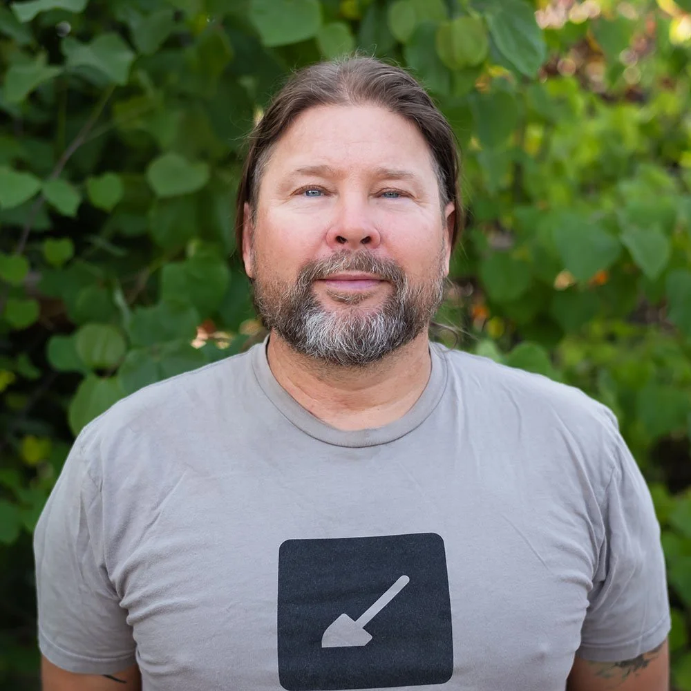 bearded caucasian male with hair pulled back wearing gray t-shirt with solid ground logo standing in front of leafy green shrubs