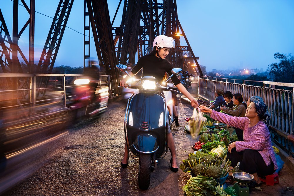 Female on scooter purchases vegetables from farmer at dusk on Long Bien Bridge, Hanoi as traffic passes.