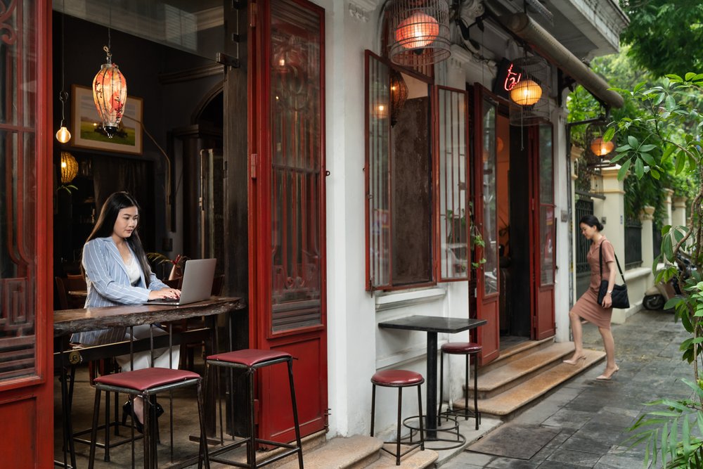  Female financial professional types on laptop at open window in artistic cafe as another woman enters cafe entrance. 