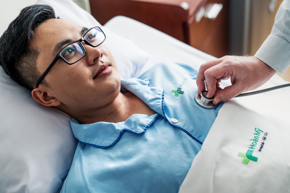 Male Patient in hospital bed as his health is checked on by nurse with stethoscope.