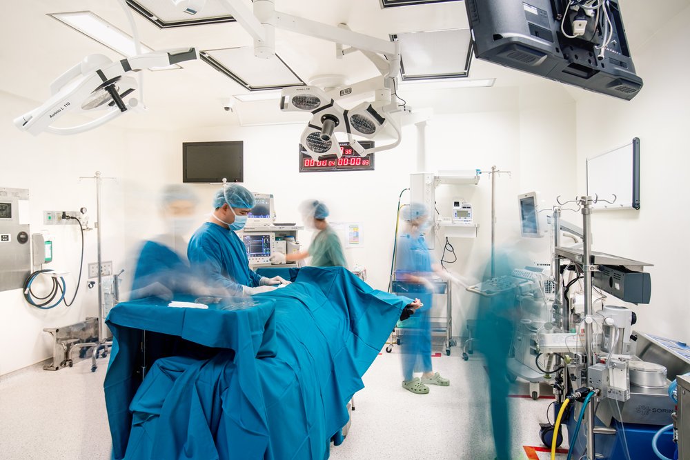 A Dr performs and operation as surgeons are blurred with motion as they move around the operating room. 