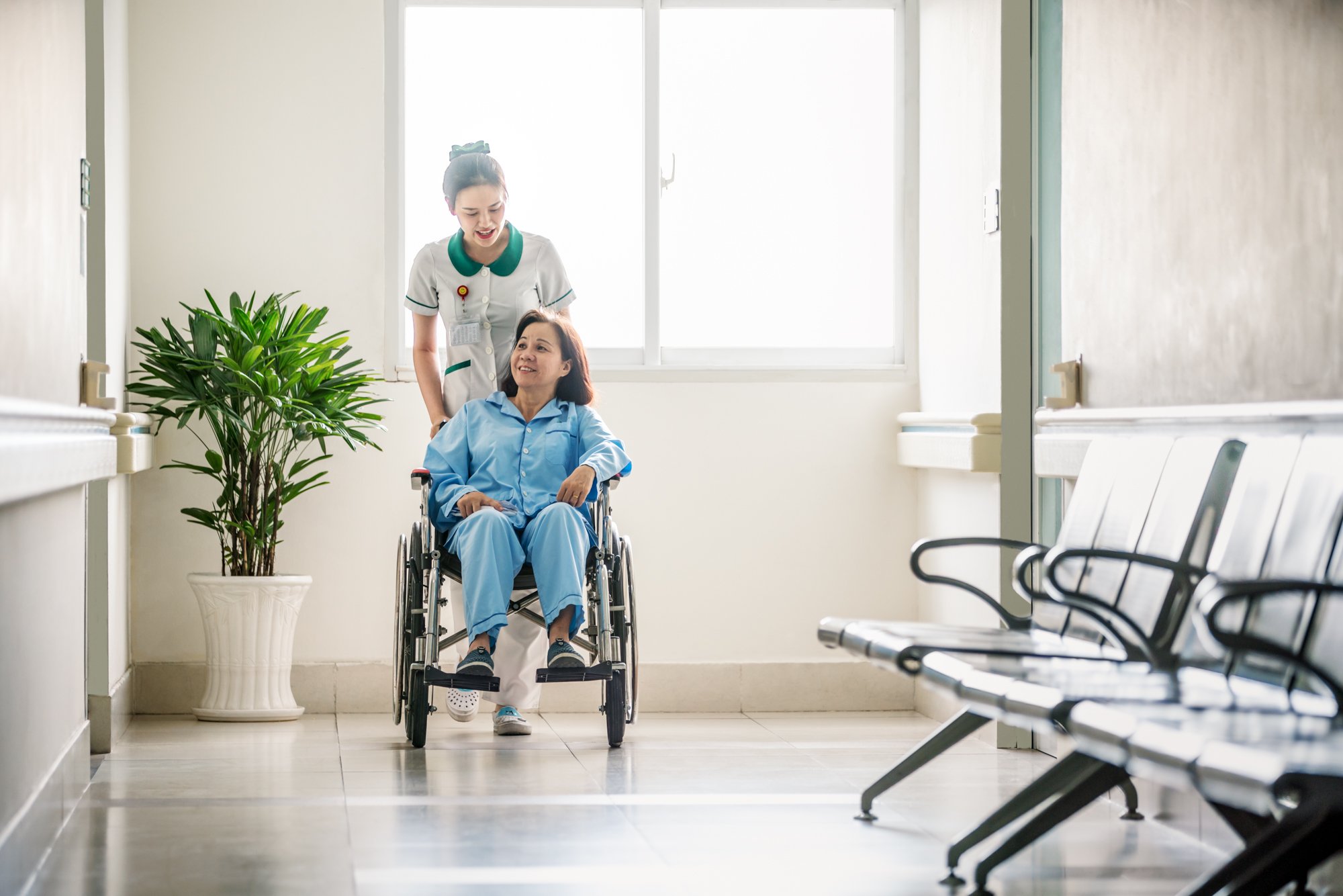 A nurse pushes and chats to wheelchair bound patient in hospital corridor. 
