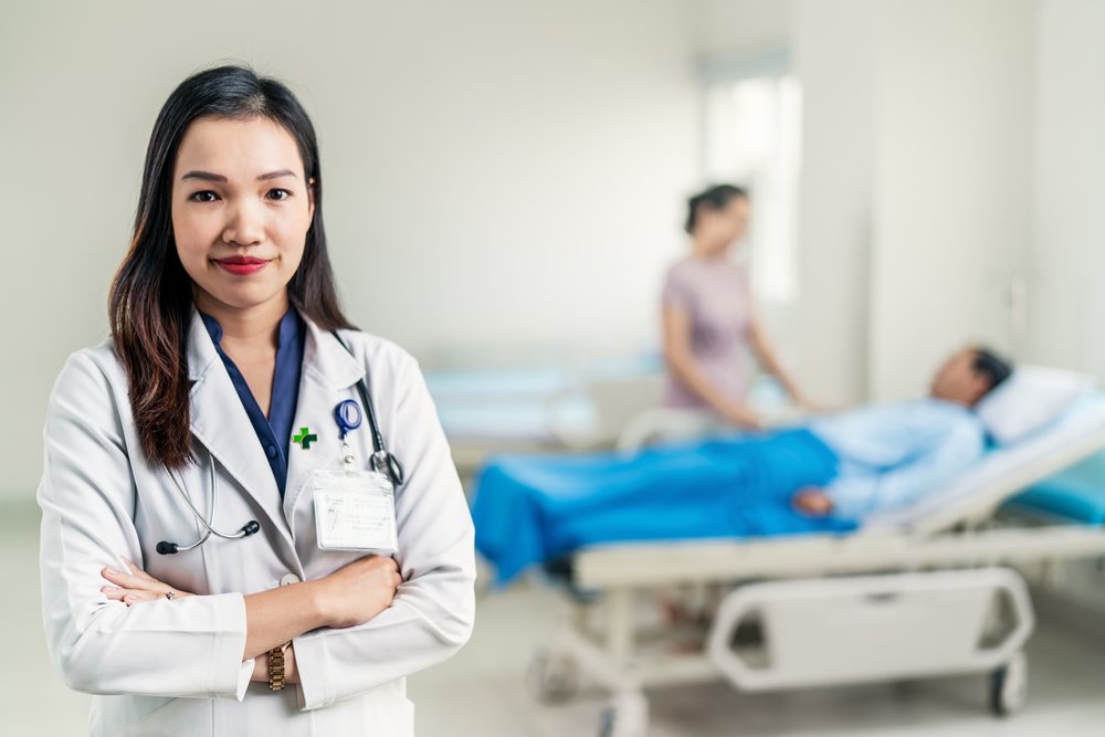 Portrait of female doctor with bed bound patient and visitor in background.