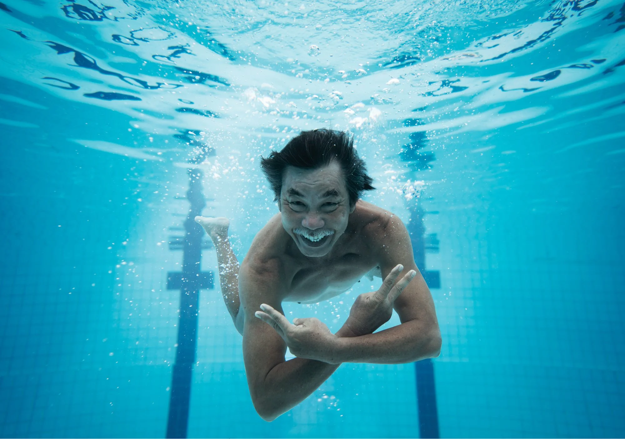  Middle aged Asian man gives V sign underwater in public pool. 