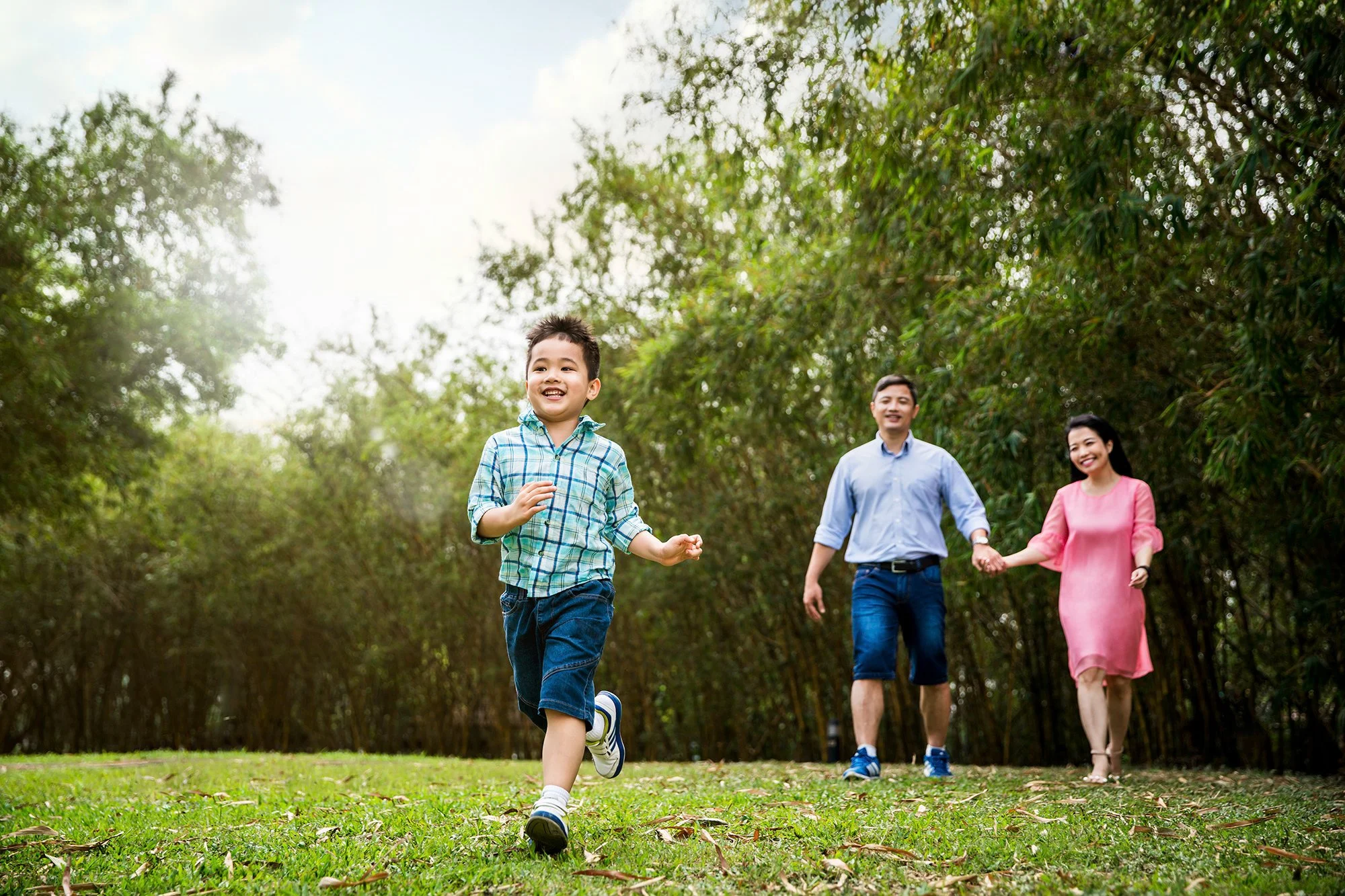  Young asian boy runs ahead of parents as they walk amongst trees in park. 